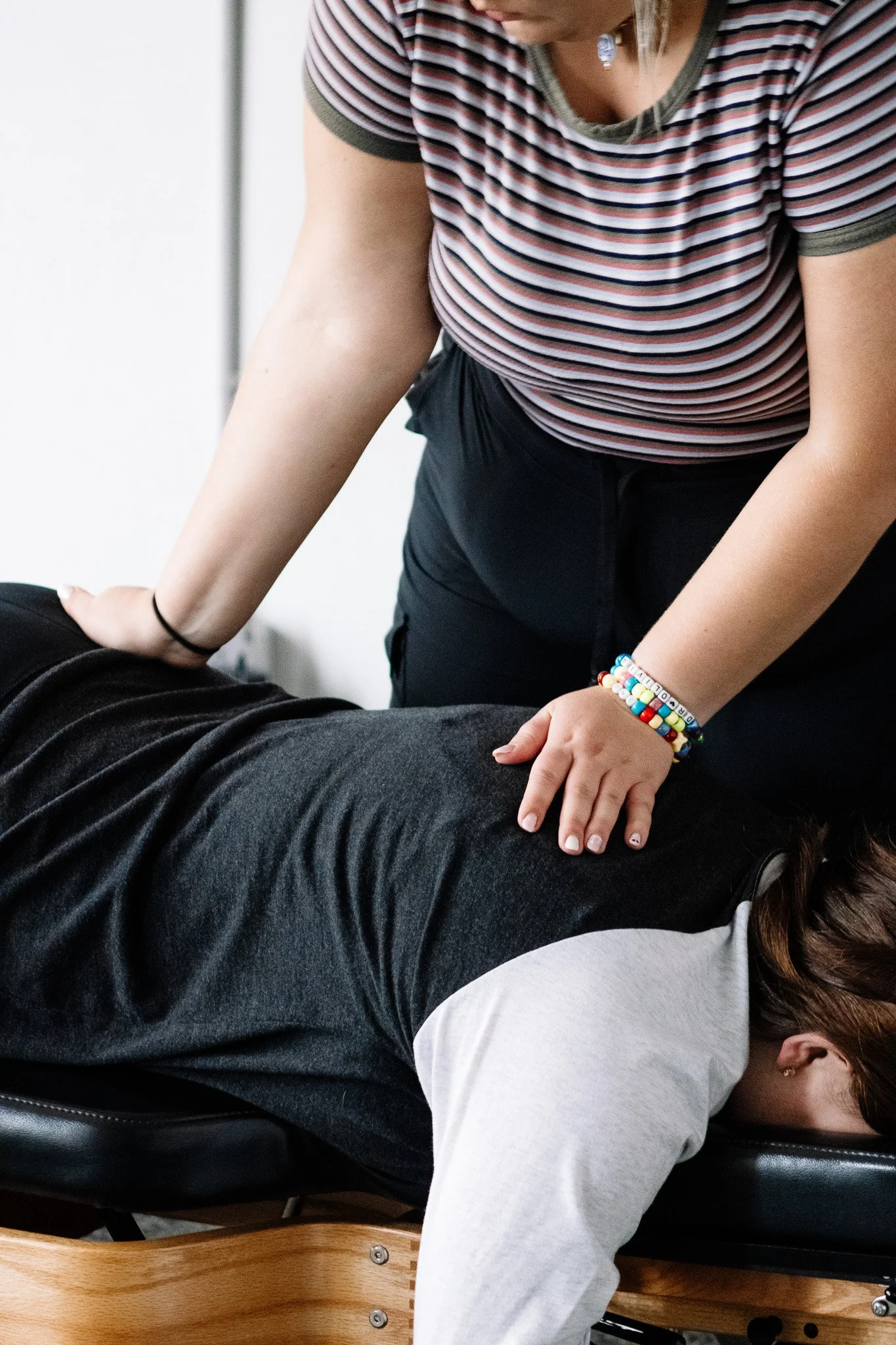 A person in a striped shirt and beaded bracelet applies pressure to a person's back during a massage. The person receiving the massage is lying face down on a massage table, wearing a gray shirt and dark pants.