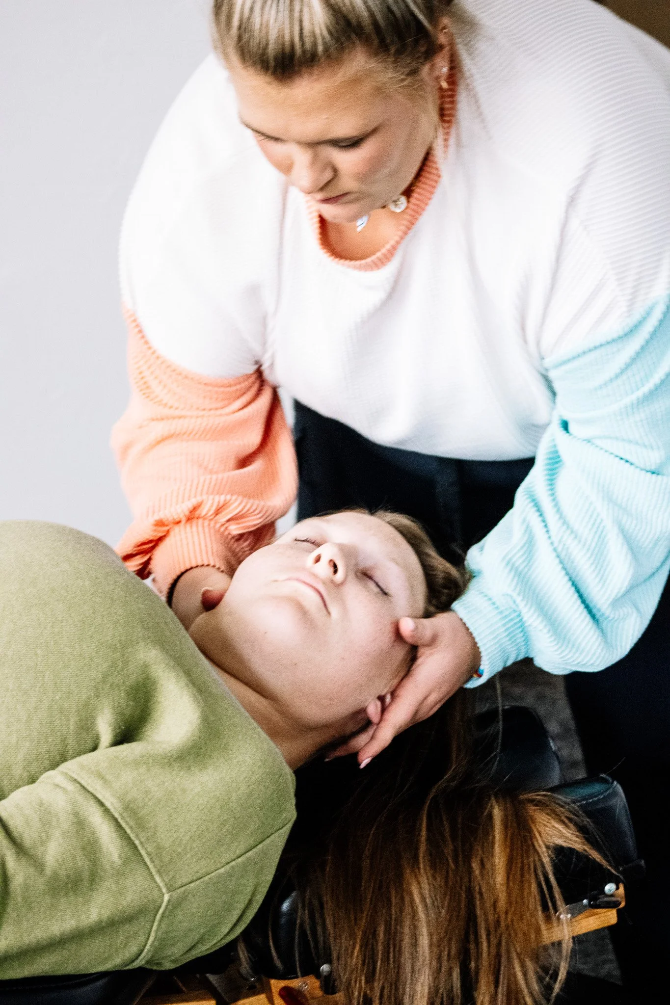 A person appears to be performing another person who is lying down with their eyes closed. The person giving aid is wearing a white and pastel-colored sweater, and has their hand on the chin of the person on the ground, who is wearing a green shirt a