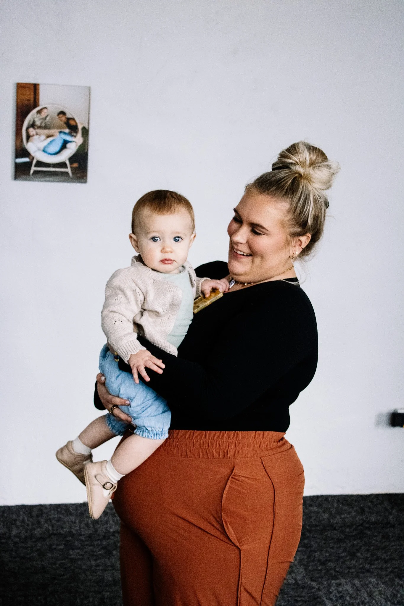 A woman with blonde hair tied up in a bun wearing a black top and orange-brown pants holds a young boy with red hair, blue eyes, and fair skin. The boy is wearing a light-colored cardigan, a white shirt, blue shorts, and beige shoes. They are standing indoors in front of a white wall with a small framed photo of two children in a modern white chair.