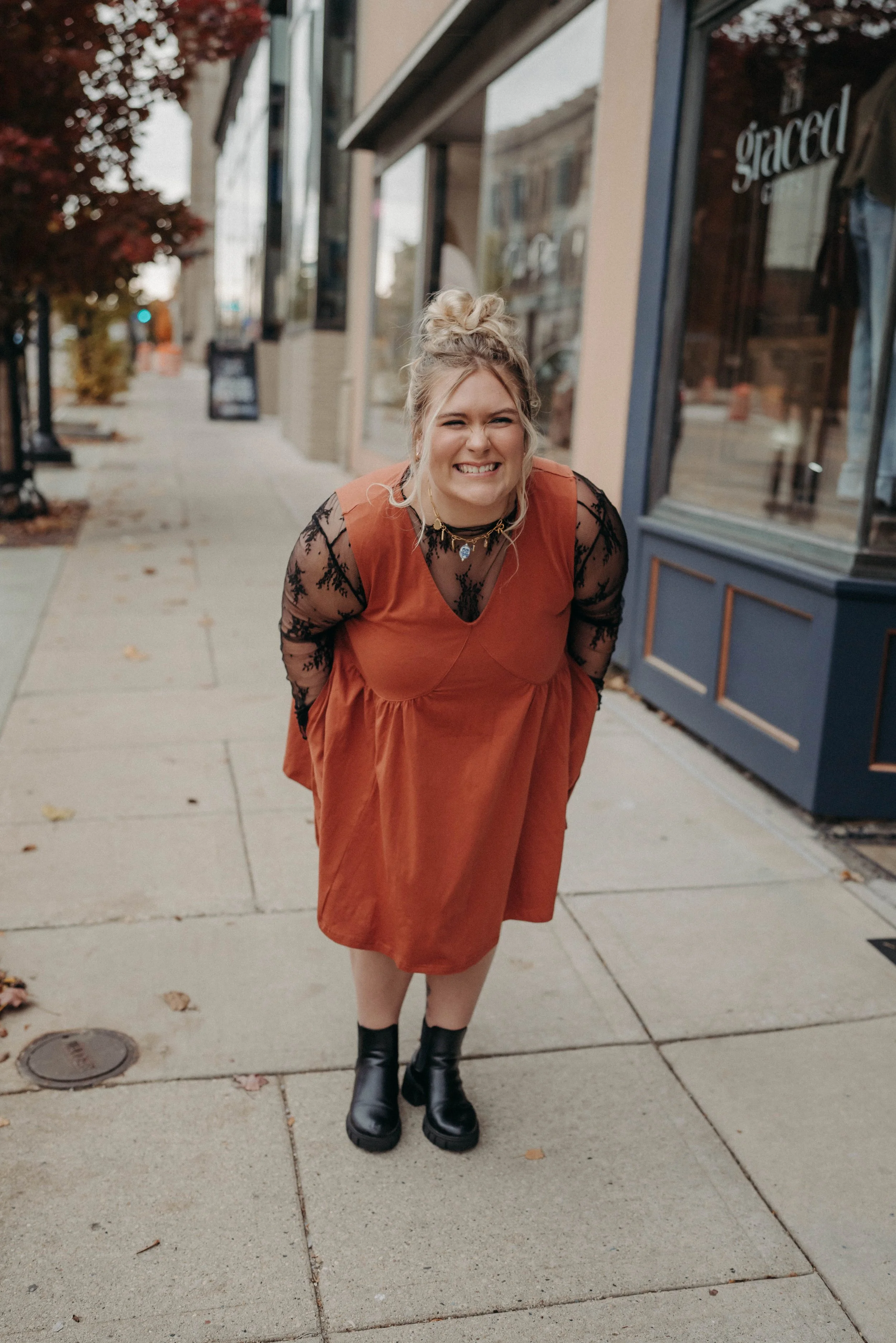 A young woman with blonde hair in a messy bun, wearing a rust-colored dress with black lace sleeves, black boots, and layered necklaces, standing on a city sidewalk, smiling with her teeth showing, in front of a storefront with a window and sidewalk trees.