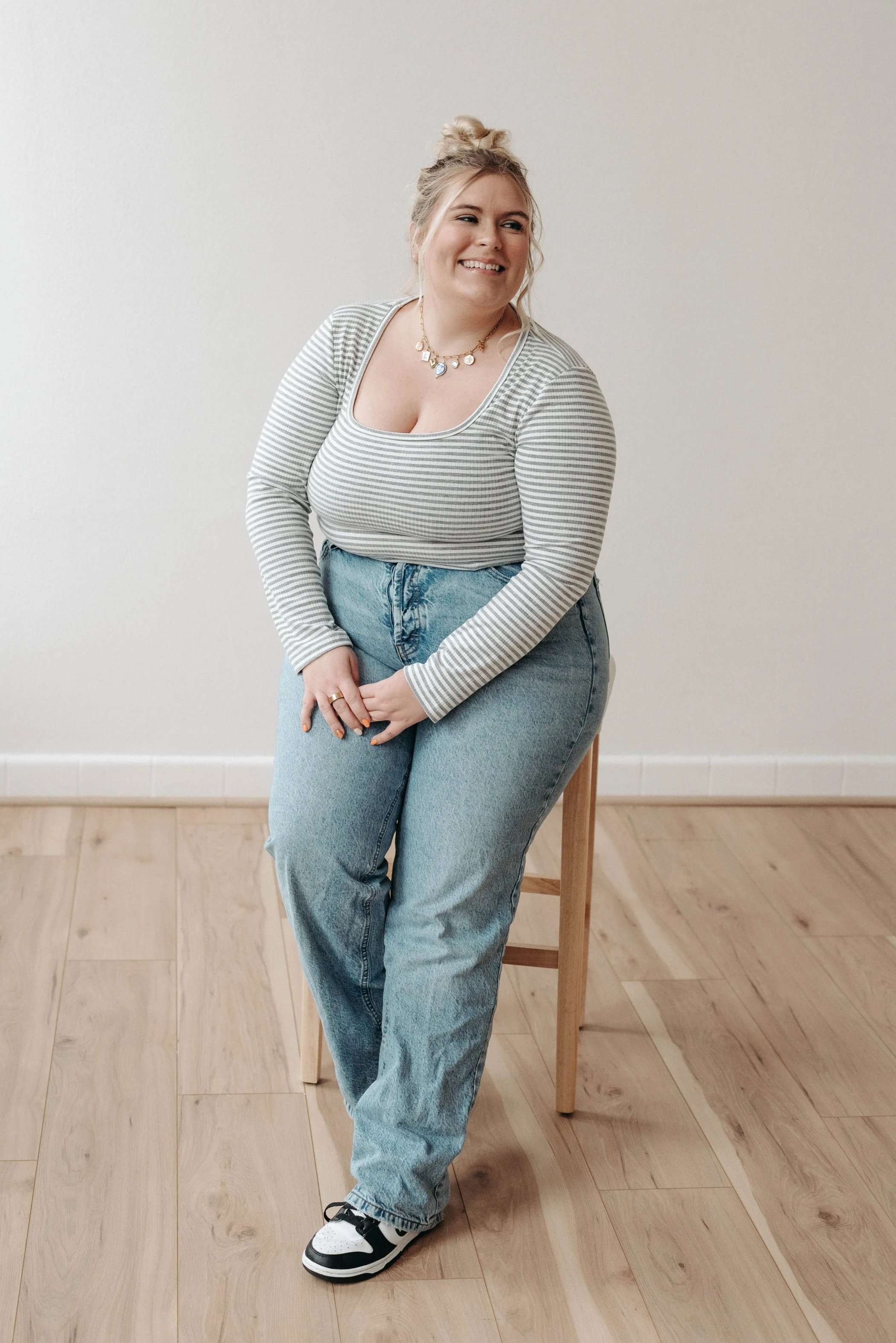 A woman with blonde hair in a messy bun, wearing a striped long-sleeve shirt, high-waisted jeans, and sneakers, sitting on a wooden stool in a room with light-colored walls and floor.