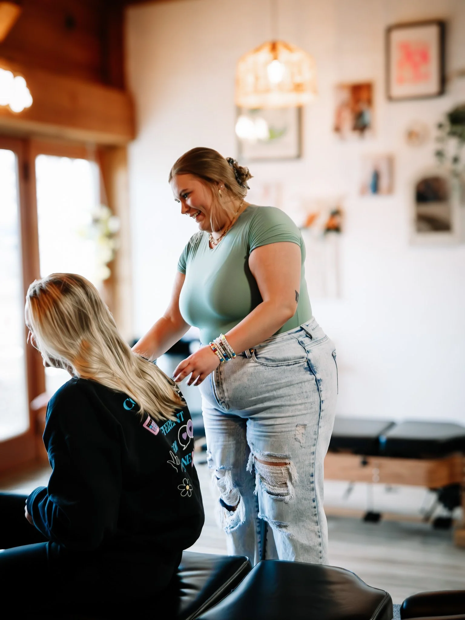 A woman with brown hair tied back, wearing a green top and ripped jeans, smiling and leaning forward while interacting with a seated blonde woman in a black hoodie, in a cozy, well-lit room with artwork and framed pictures on the wall.