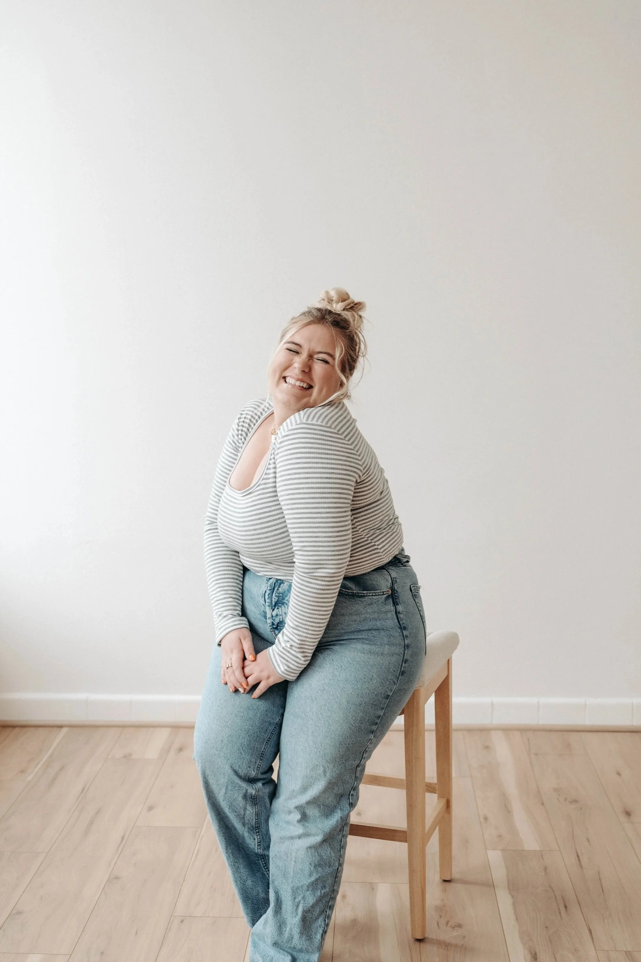 A young woman with blonde hair in a messy bun, wearing a striped long-sleeve shirt and light blue jeans, sitting on a wooden stool against a plain white wall, smiling with eyes closed.