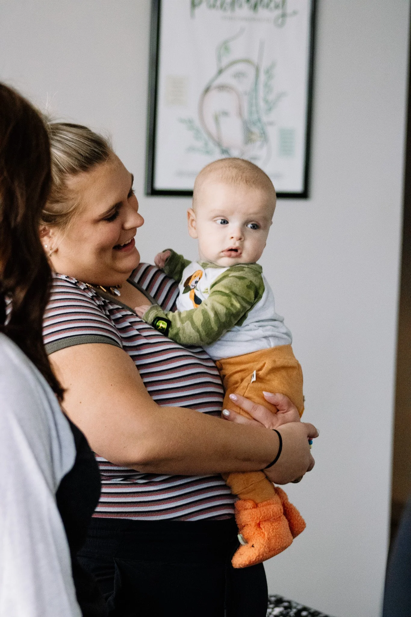 A smiling woman holding a baby with blonde hair and blue eyes, dressed in a hoodie with animal print, appears to be in a medical or professional setting, with a diagram in the background.