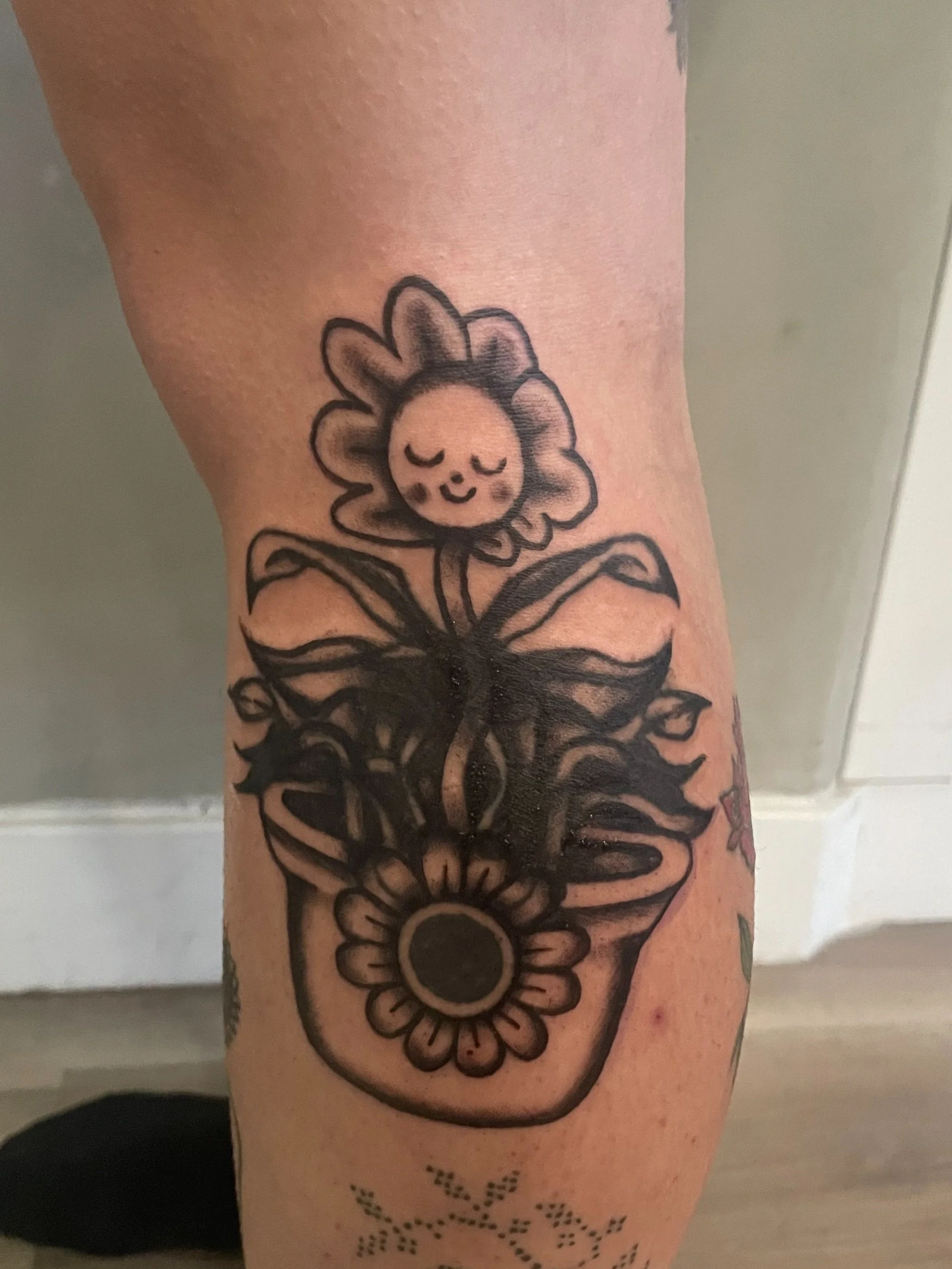 Black outline tattoo of a potted flower with a smiling face, surrounded by leaves and a sunflower at the base, on a person's skin.