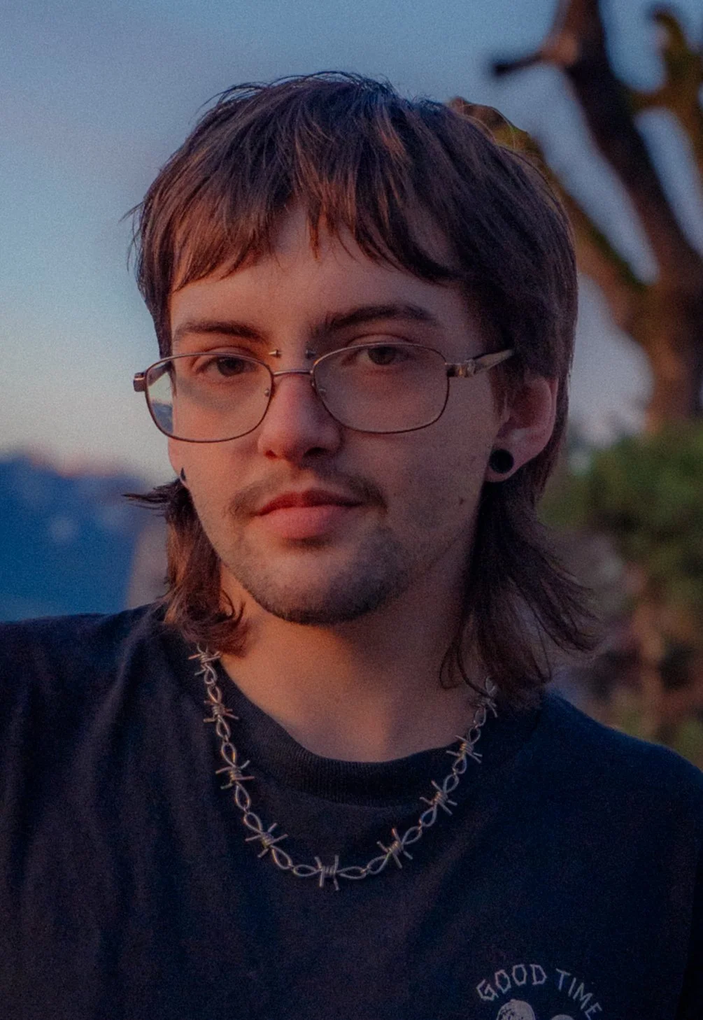 A young person with glasses, dark brown hair, and a chain necklace, outdoors during sunset, wearing a black shirt with partially visible white text.