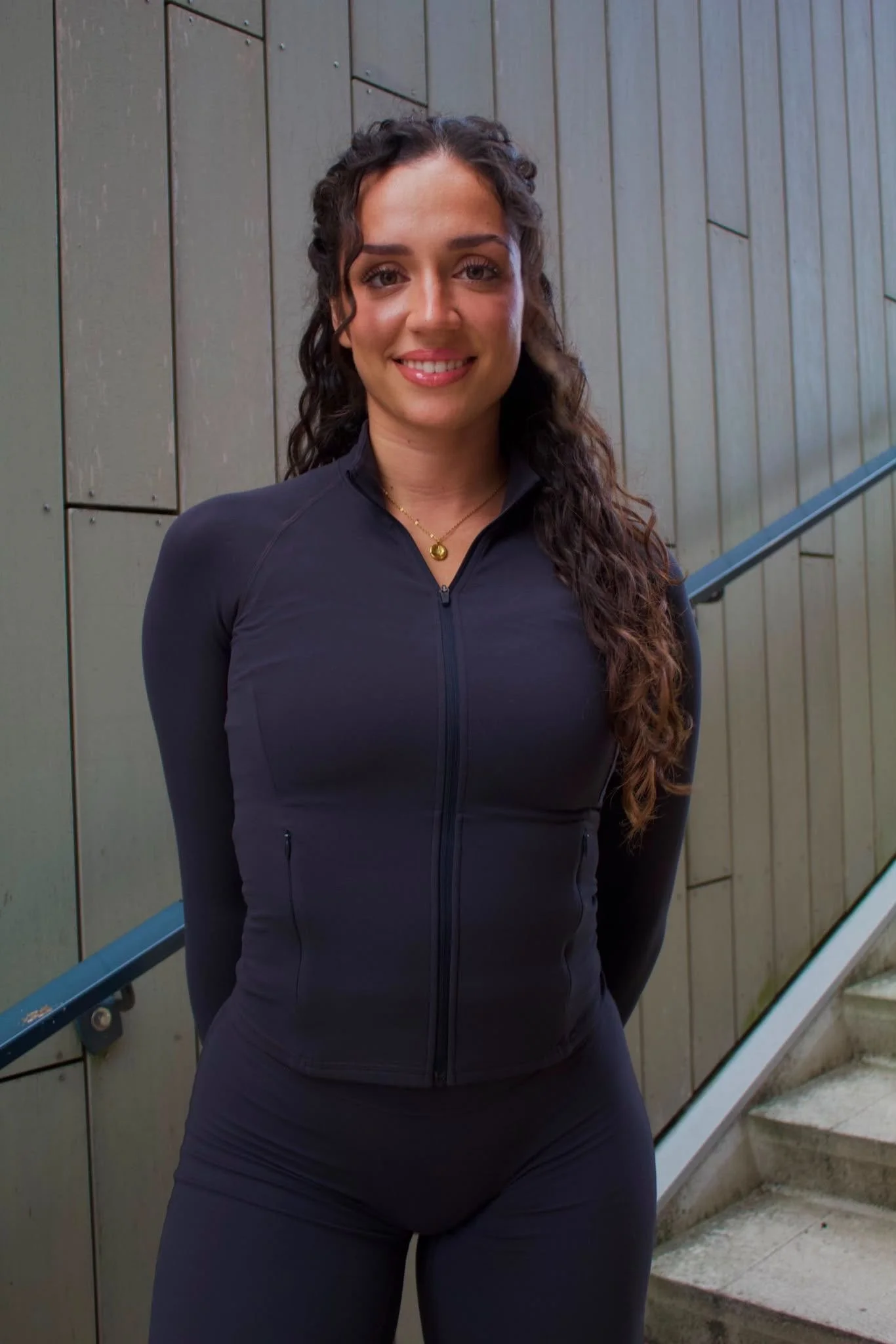 Een vrouw met lang, golvend haar, gekleed in een zwart sportoutfit, staat buiten voor een betonnen muur met trap. Ze glimlacht naar de camera.