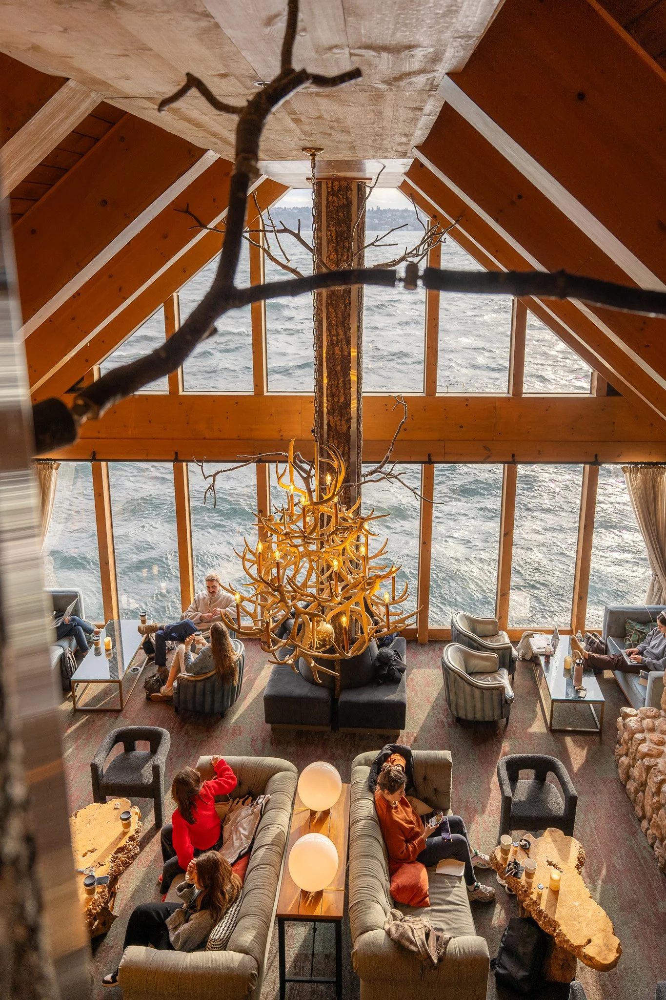 Interior of a cozy lounge with large windows overlooking water, featuring a large antler chandelier, a central tree trunk with branches, and several people sitting on sofas and chairs, some using phones and computers.