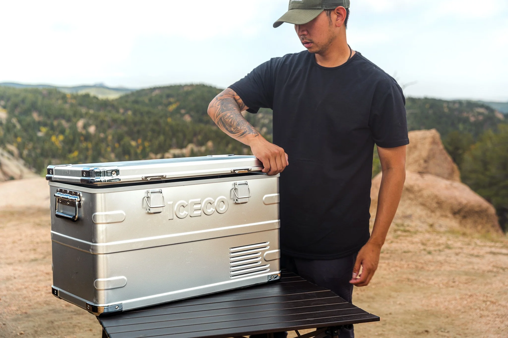 A man wearing a gray baseball cap and a black t-shirt is outdoors, opening a silver portable refrigerator with the brand name "ICECO." The background features hills and trees under a cloudy sky.