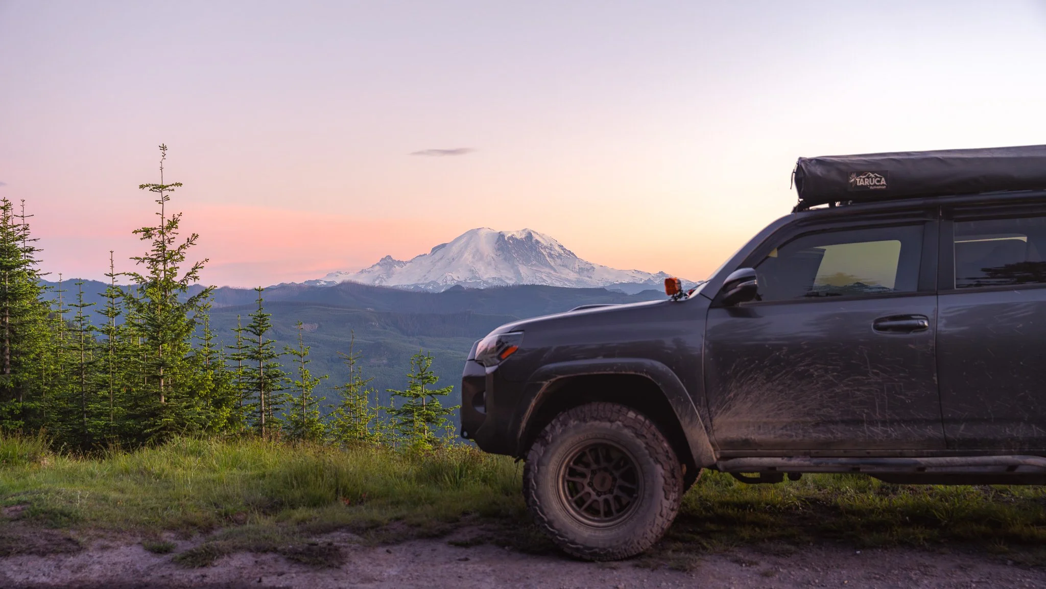 A black off-road vehicle with a rooftop tent parked on grassy area with trees, with snow-capped mountain in the background during sunset.