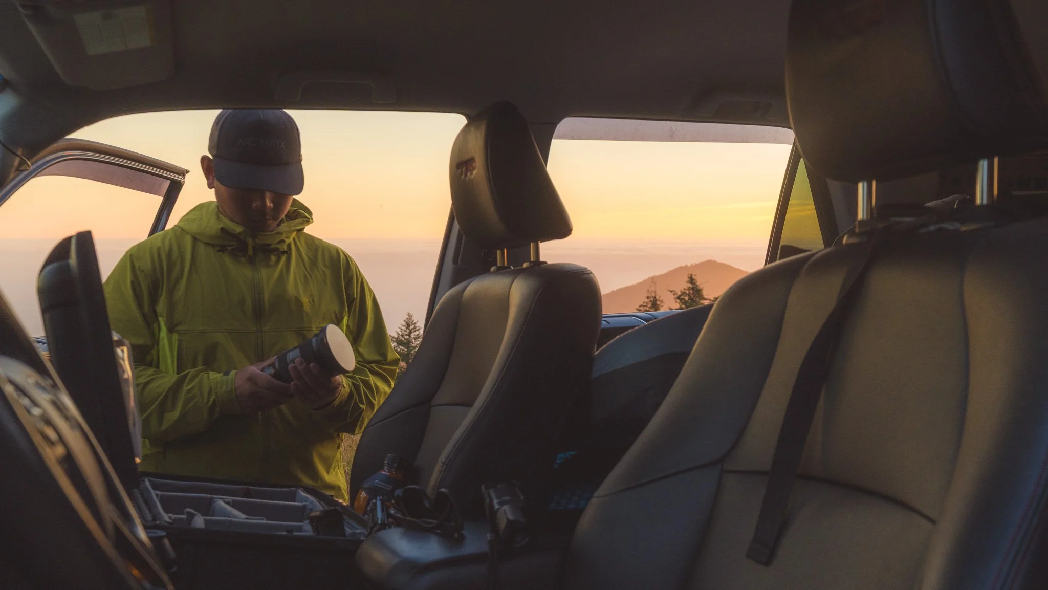 A person wearing a yellow jacket and a dark cap standing inside a vehicle at sunset, holding a flashlight or small device, with mountain and sky background visible through the vehicle windows.