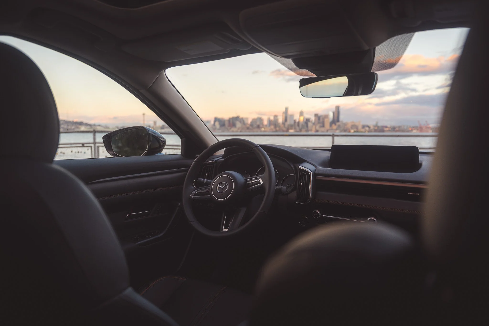 View of a city skyline at sunset from inside a car parked near a body of water, with a blurry dashboard and steering wheel in the foreground.