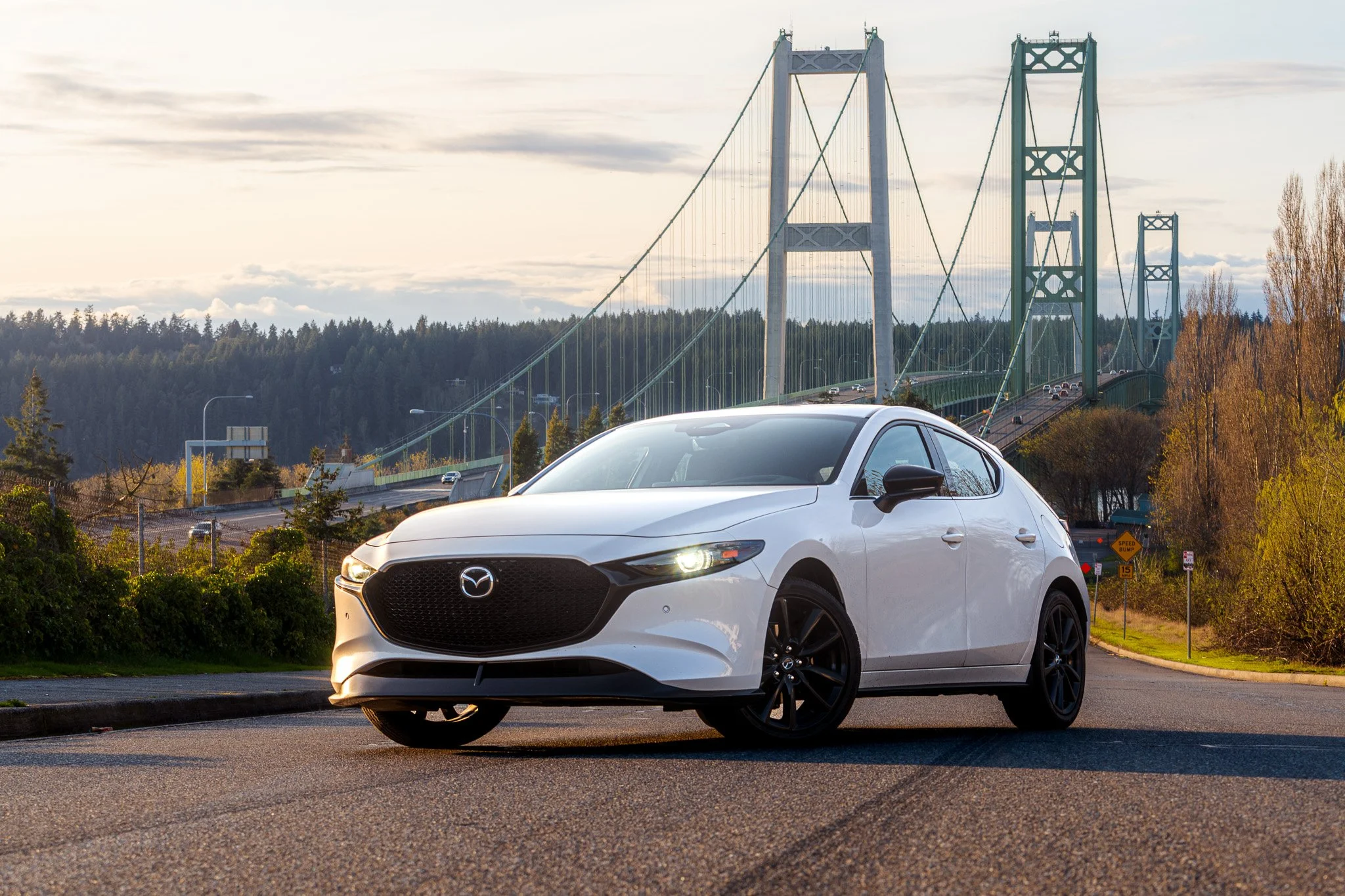 White Mazda car parked on the side of a road near a bridge, with trees and a cloudy sky in the background.