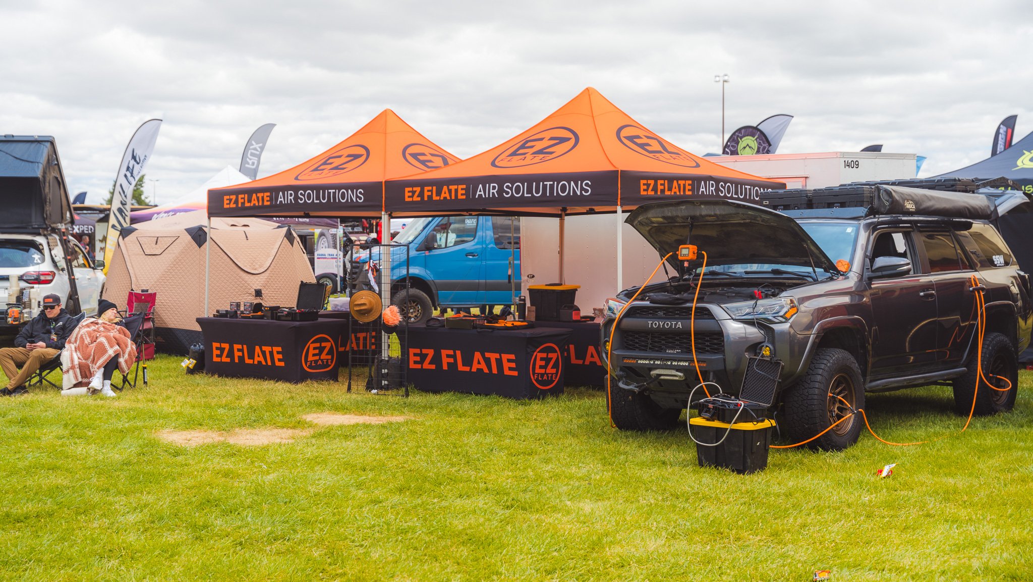 An outdoor trade show with tents and booths, including one with a sign for EZ Flate Air Solutions. There are vehicles, camping gear, and people sitting and walking on the grass field under a cloudy sky.
