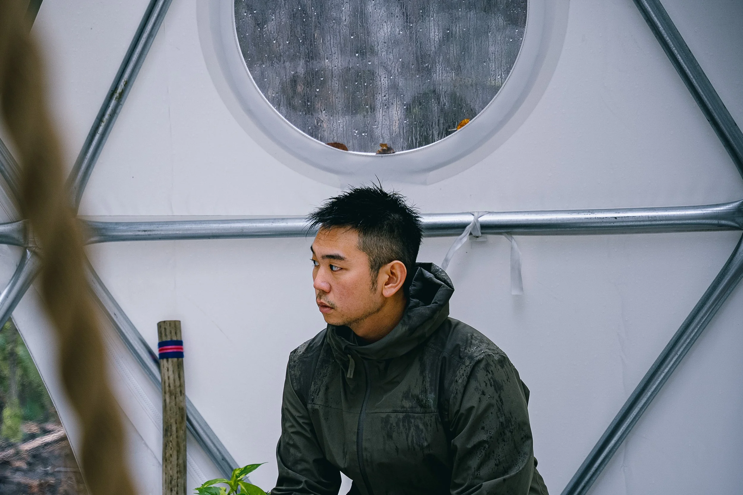 A young man with short black hair and a dark jacket looking to his left, standing inside a greenhouse with a round window and metallic framing.