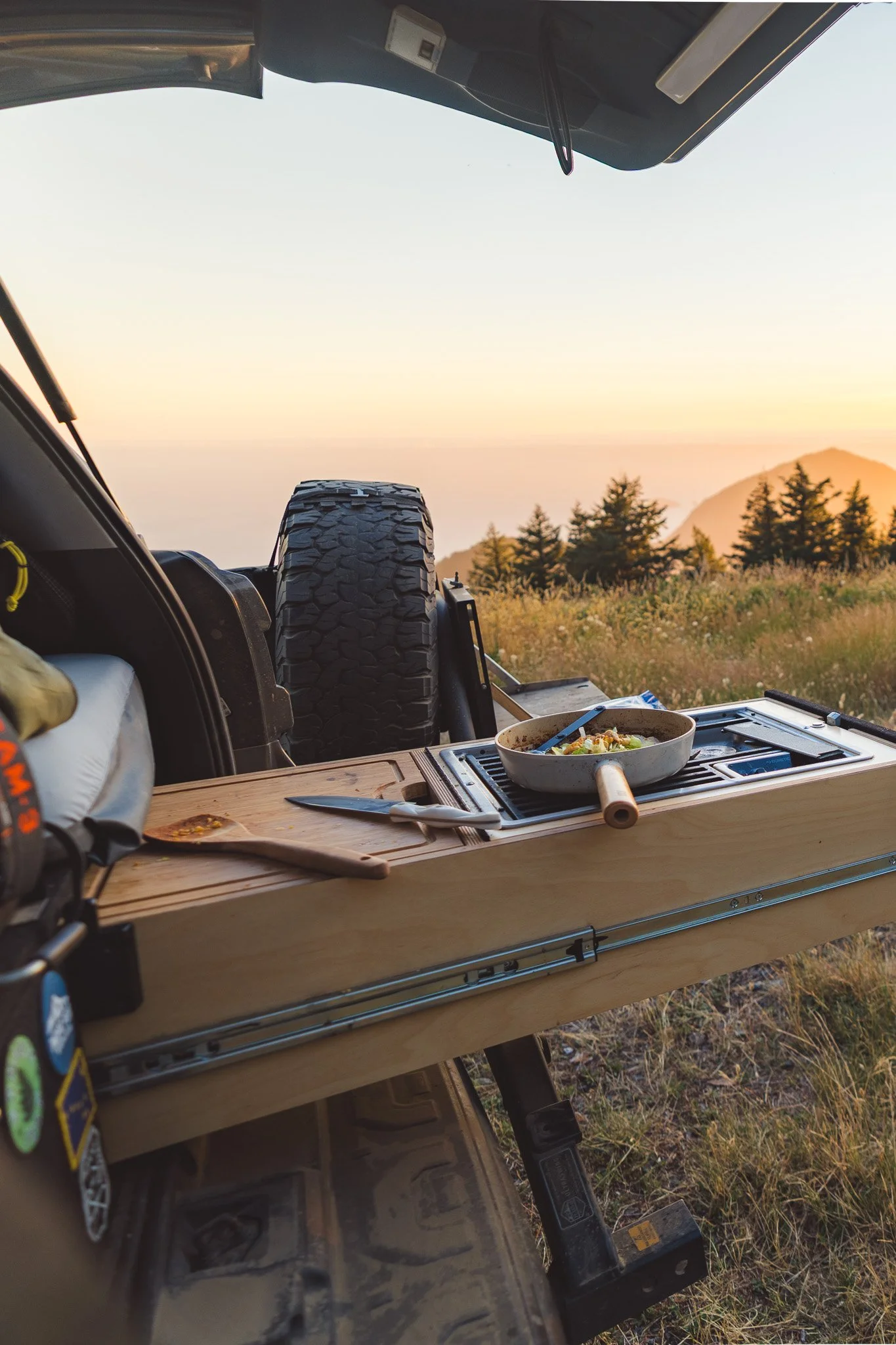 A view from inside a vehicle with a spare tire, showing an outdoor scene during sunset with trees and mountains in the background. There is a wooden table with a skillet containing food, a knife, and a cutting board with some food on it.