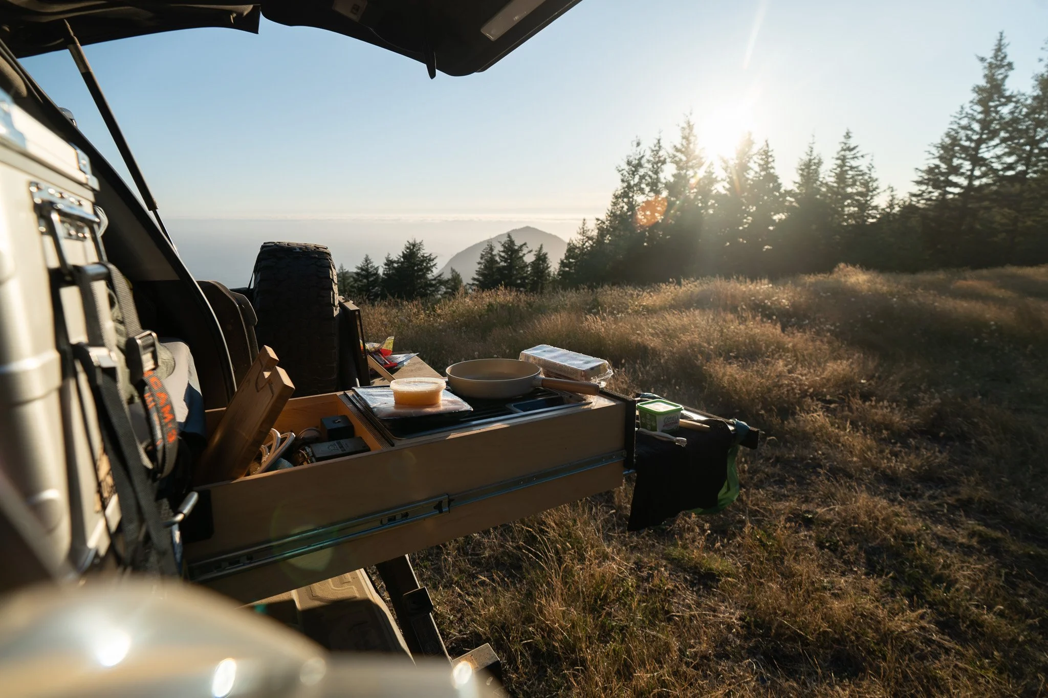 Camping scene with a table of food and outdoor supplies in a forested area during sunrise or sunset with trees and a mountain in the background.