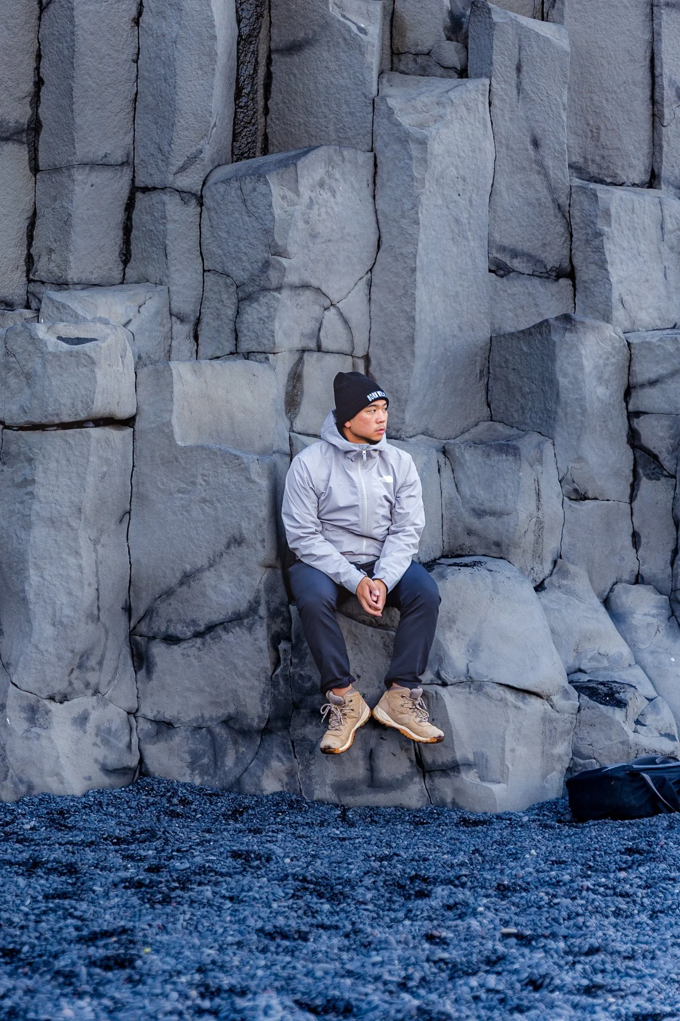 A man in outdoor clothing, including a gray jacket, beige hiking boots, and a black beanie, sitting on a rock ledge against a large stone wall with dark rocks underneath.