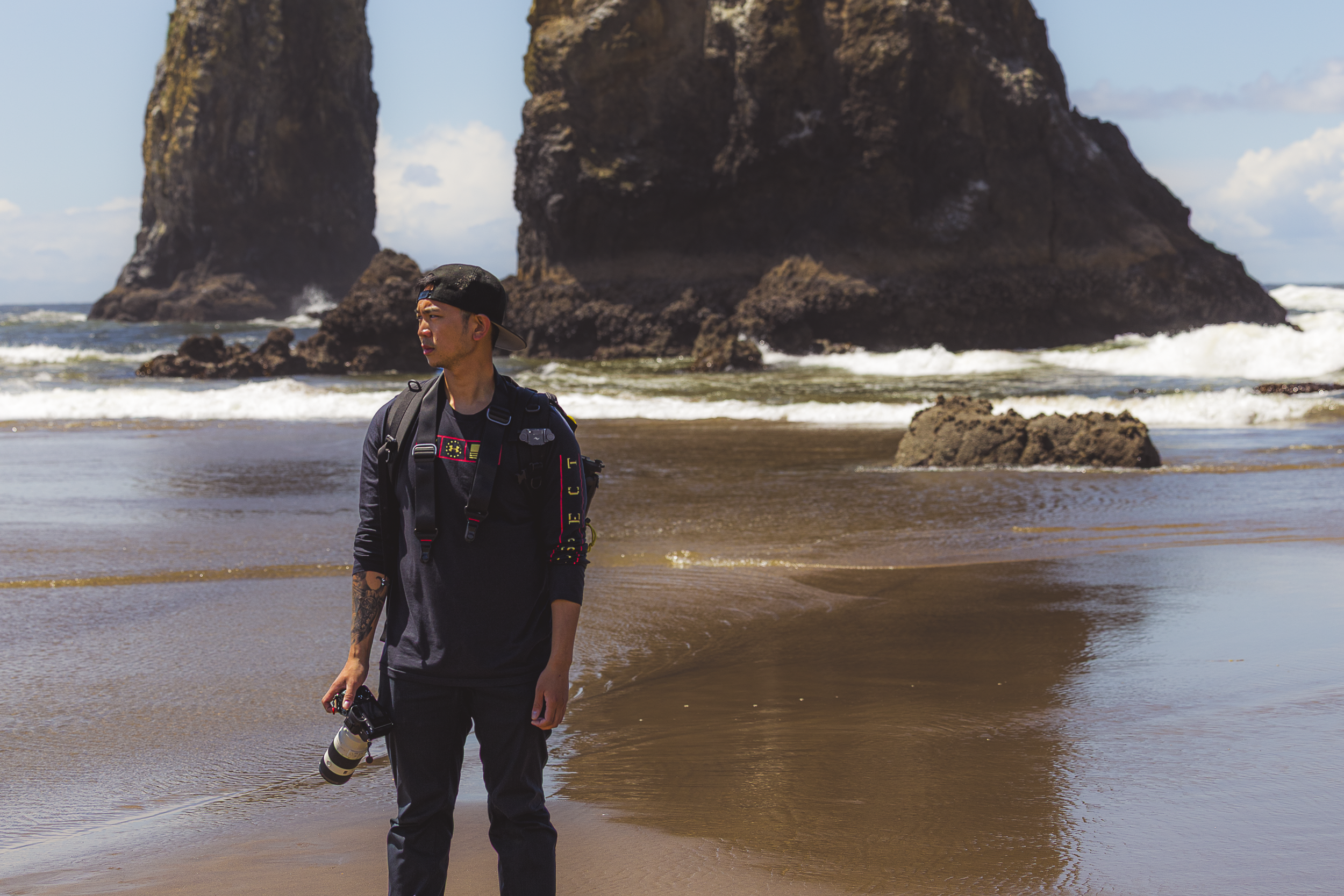 A man walking along a sandy beach holding a camera with a long lens. The man is wearing a backwards cap, black clothing, and a backpack. Large rock formations are visible in the background under a partly cloudy sky.