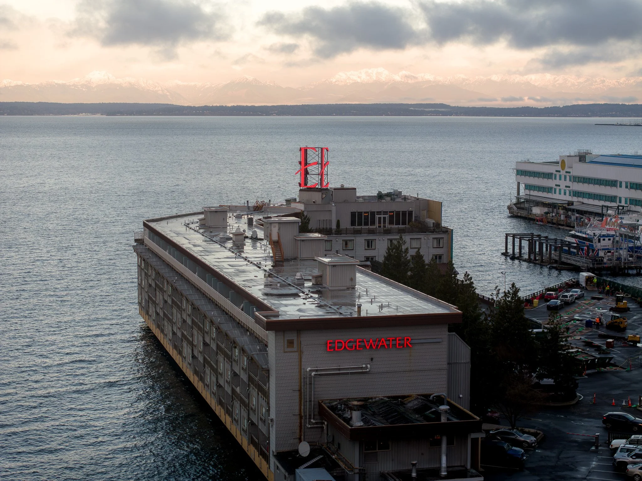 A waterfront city view featuring a large building with the word 'EDGEWATER' illuminated in red on its side, adjacent to a dock with boats, and a distant mountain range with snow-capped peaks under a cloudy sky.