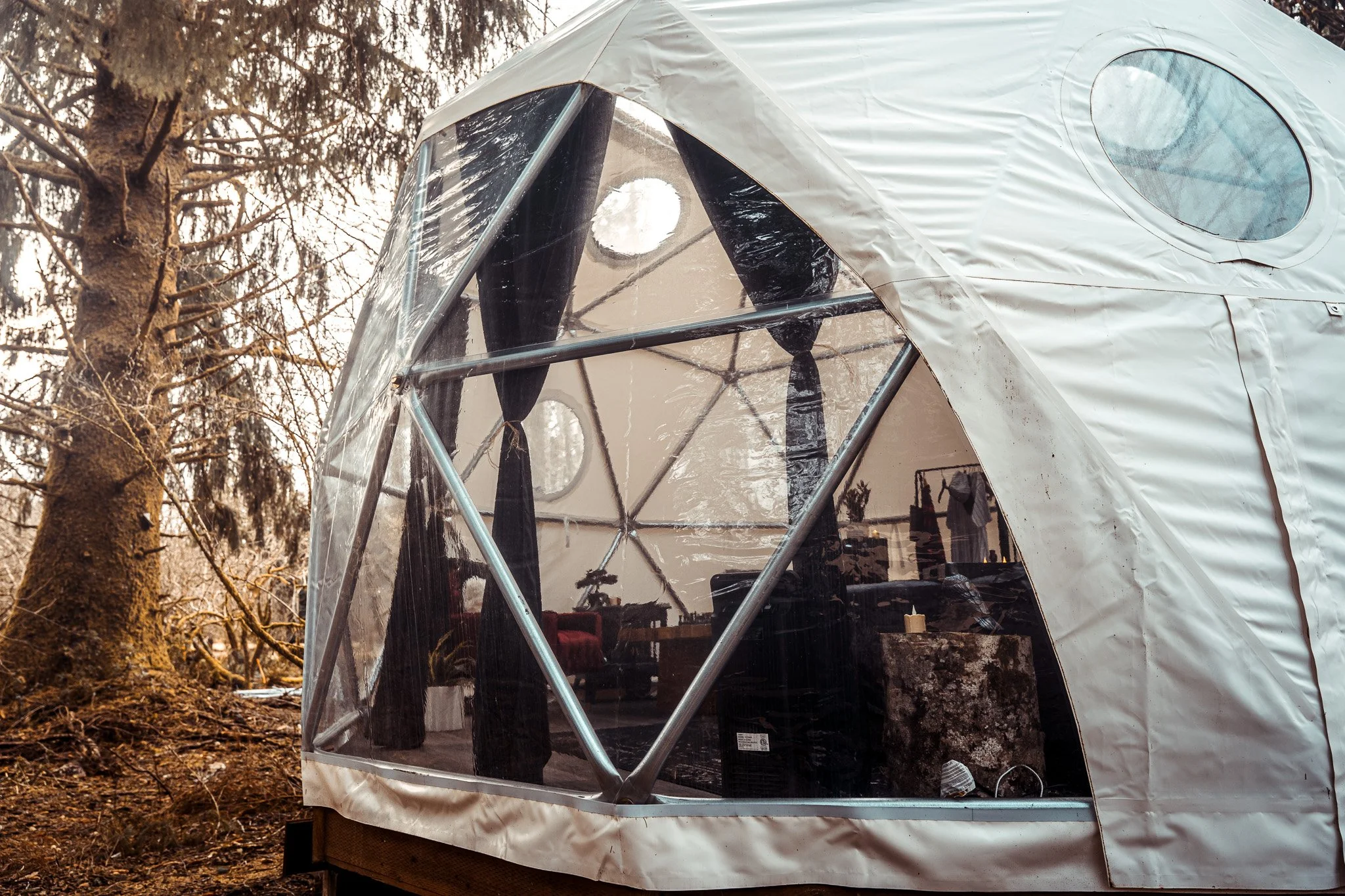 A white outdoor tent with clear plastic windows, set up in a wooded area during winter or fall.