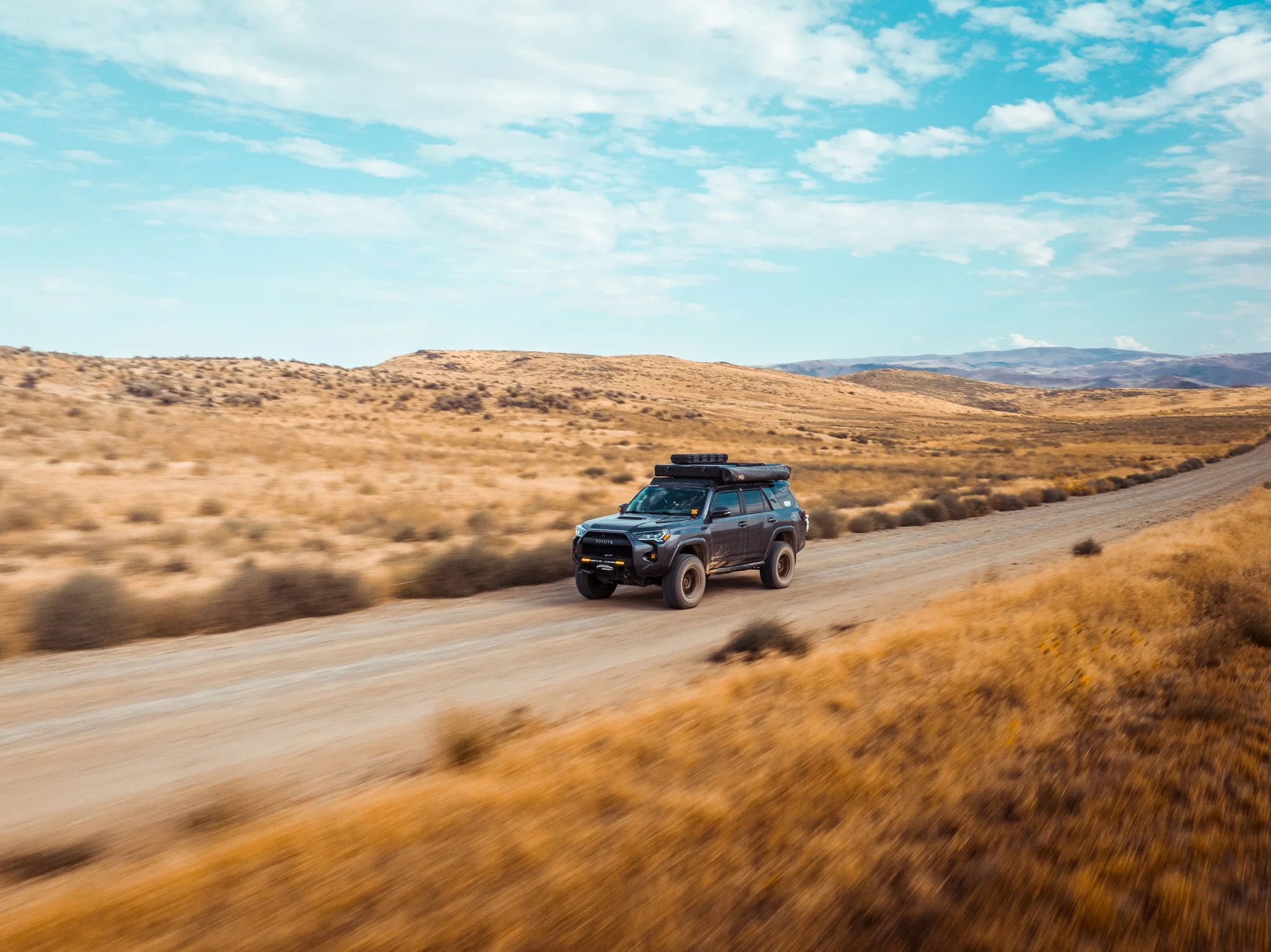 A black SUV driving on a dirt road through an arid landscape with dry grass and rolling hills under a blue sky with scattered clouds.