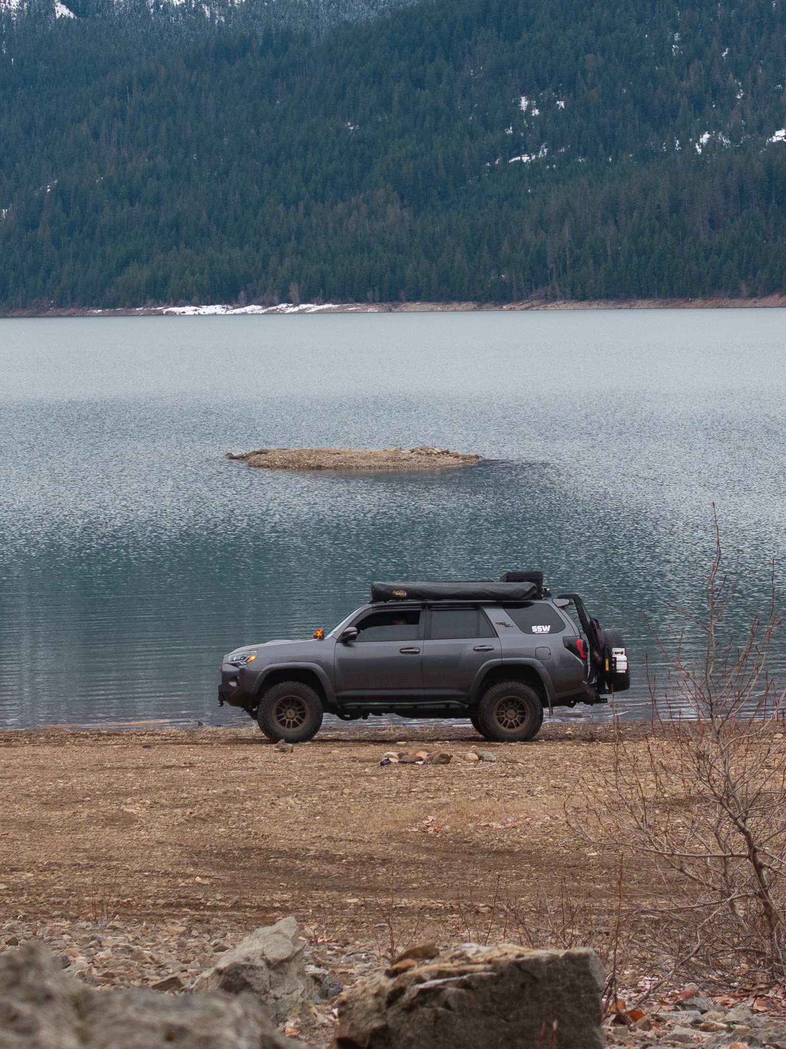 A black SUV parked near a rocky shore by a large calm lake, with a forested mountain in the background.