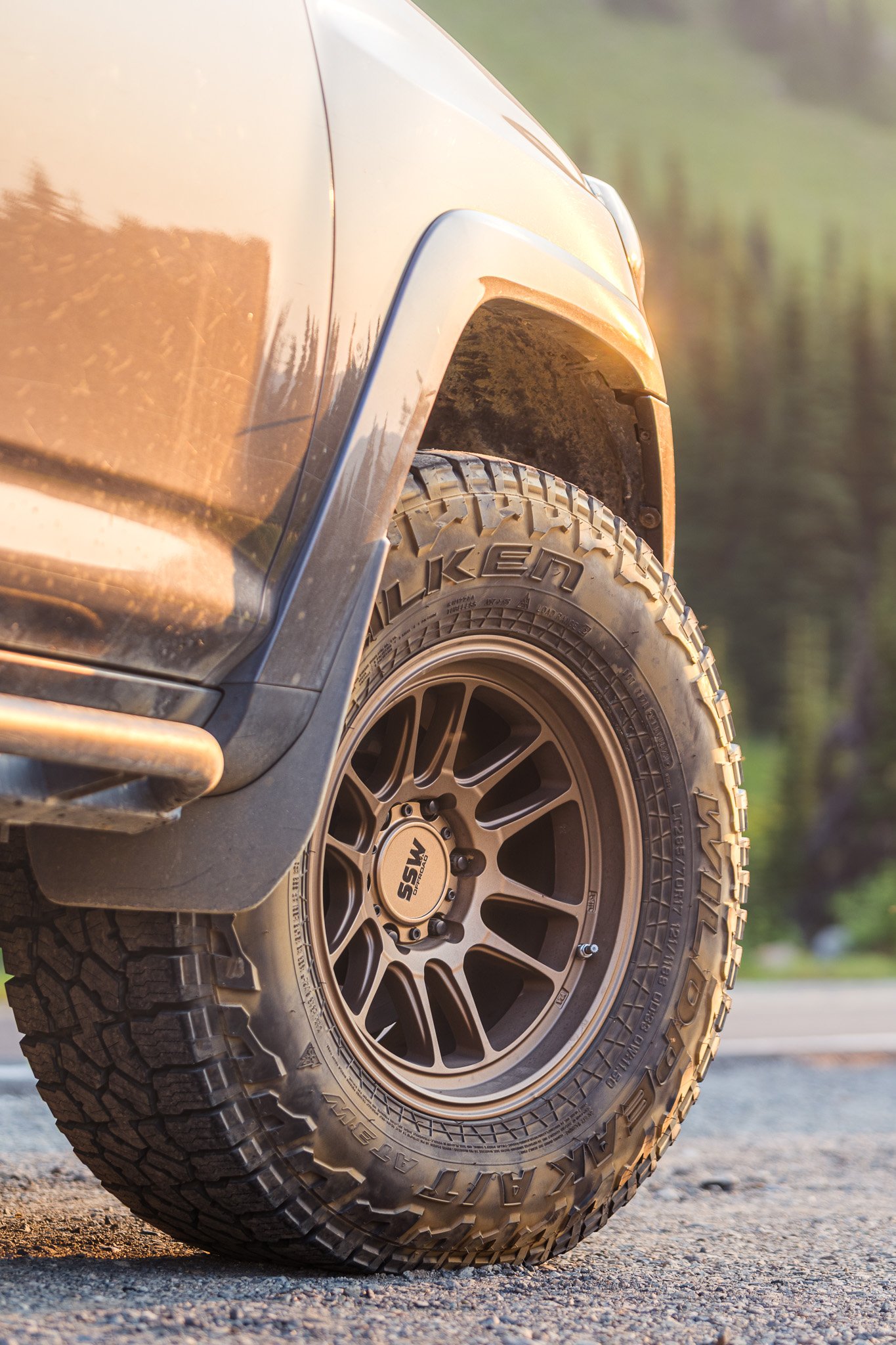 Close-up of an off-road vehicle's front wheel and tire on rocky terrain, with the vehicle's tan fender and rugged terrain in the background.