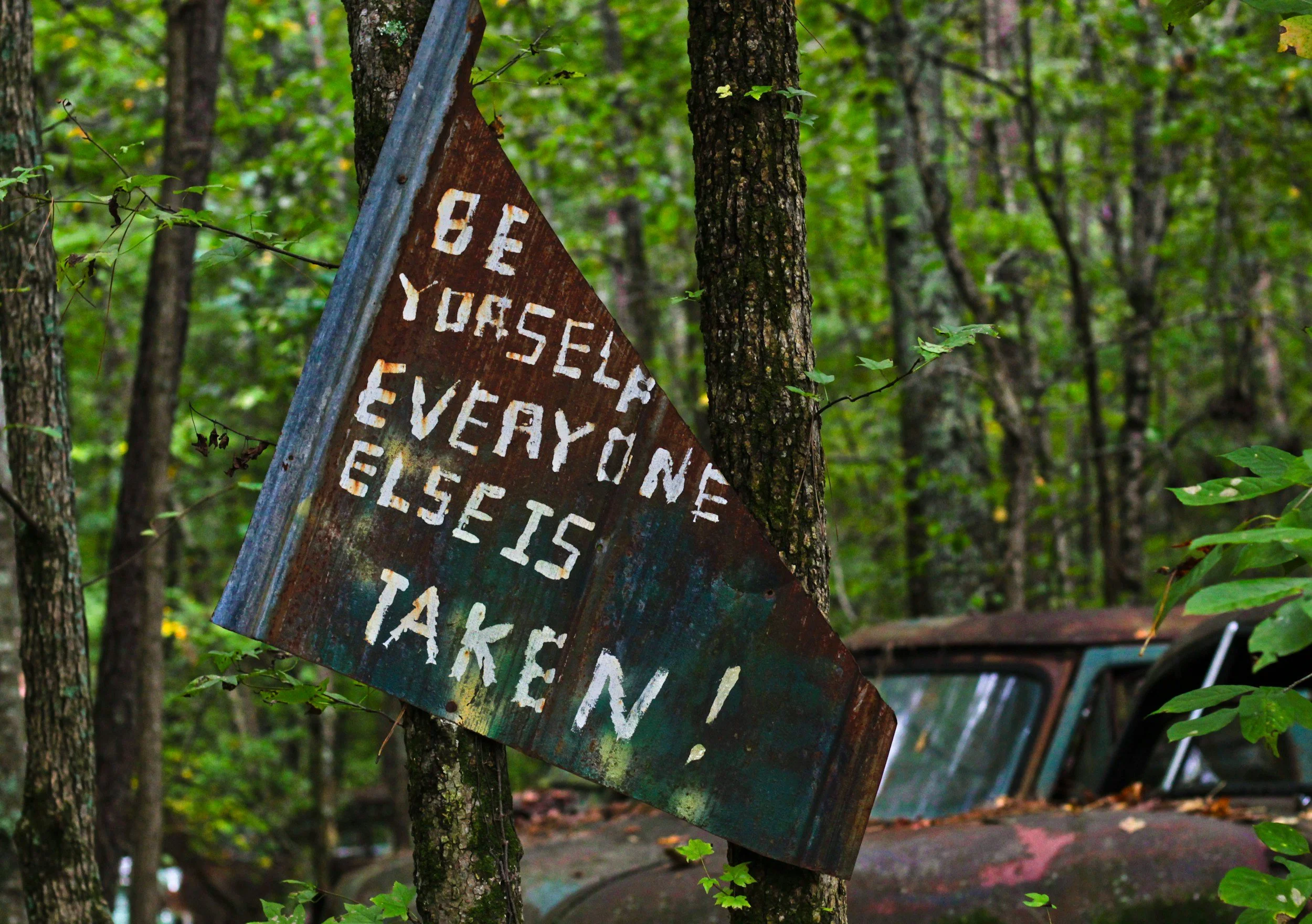 Rusty metal sign attached to a tree with a forest background, featuring partially visible white painted text.