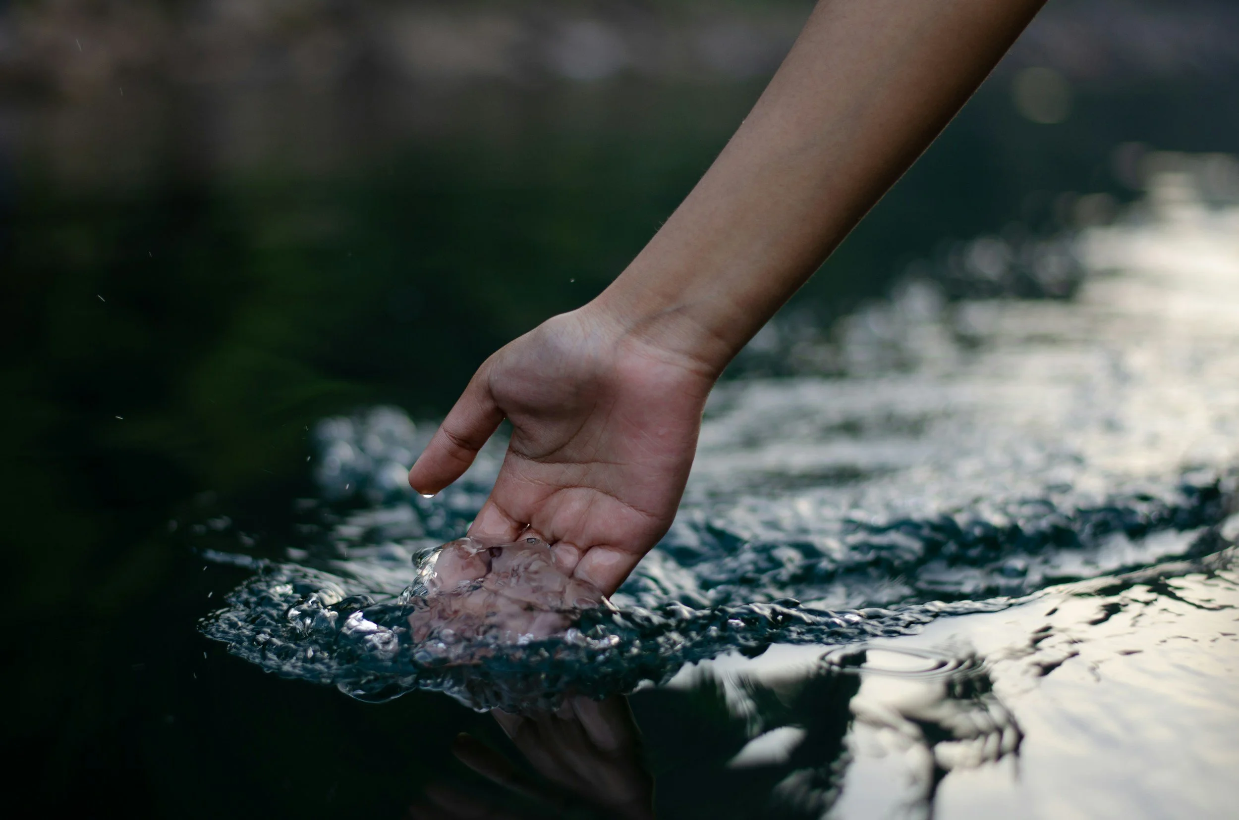 A person's hand dips into a body of water, creating ripples and small splashes on the surface.