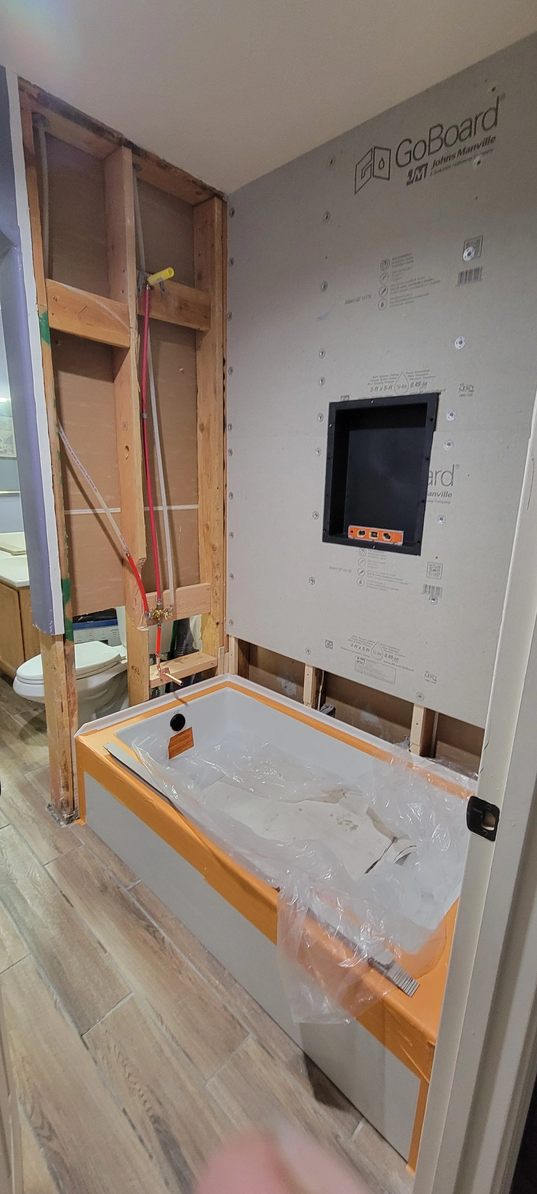 Bathroom remodel in progress, showing a bathtub with protective plastic, a wall with unfinished materials, and plumbing fixtures.