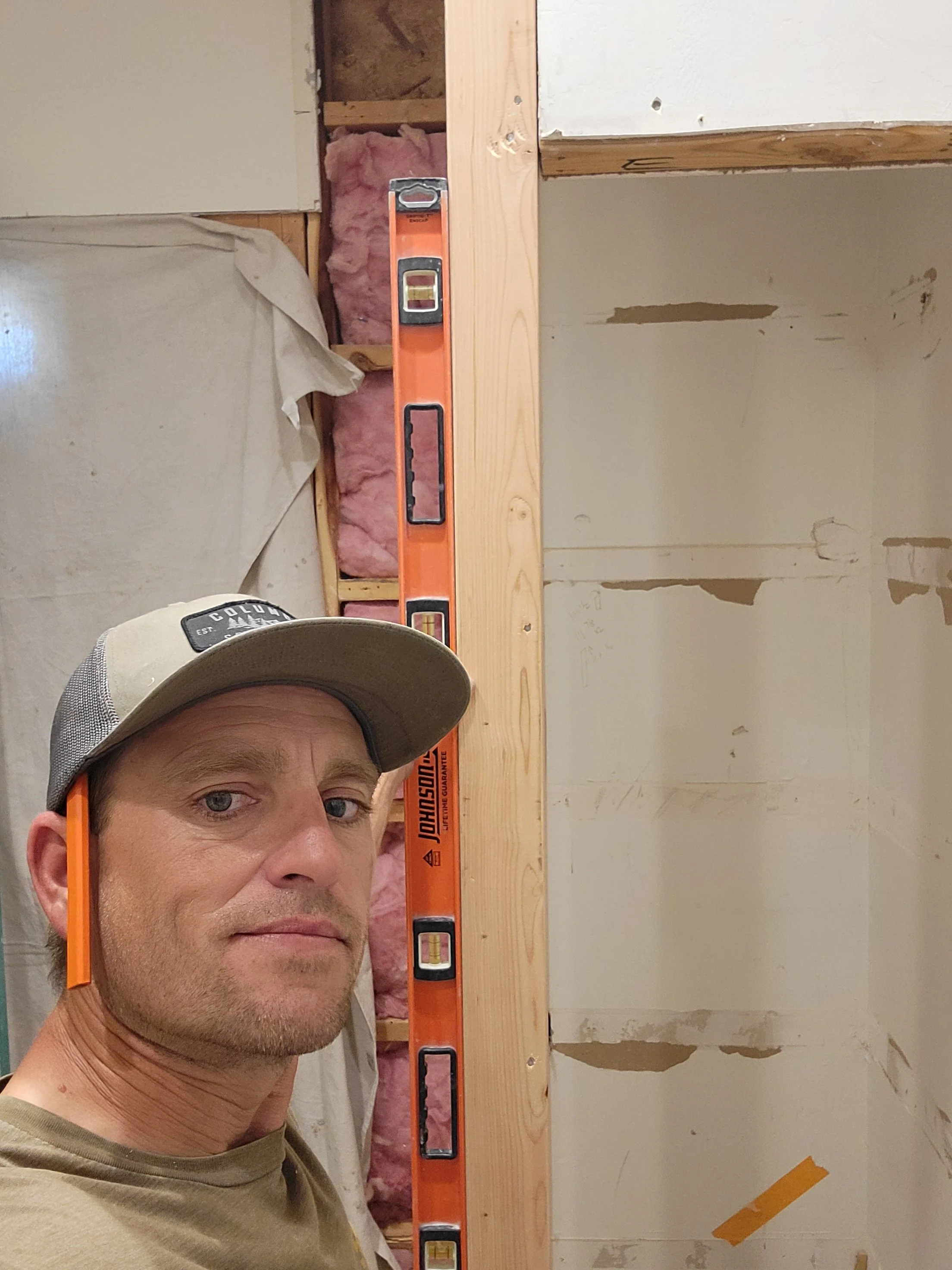 A man wearing a beige cap and green shirt taking a selfie in a room under renovation. His face appears serious or concentrated. The background shows unfinished walls with exposed pink insulation, a piece of drywall, and a wooden stud. A level tool is positioned vertically against the wooden stud.