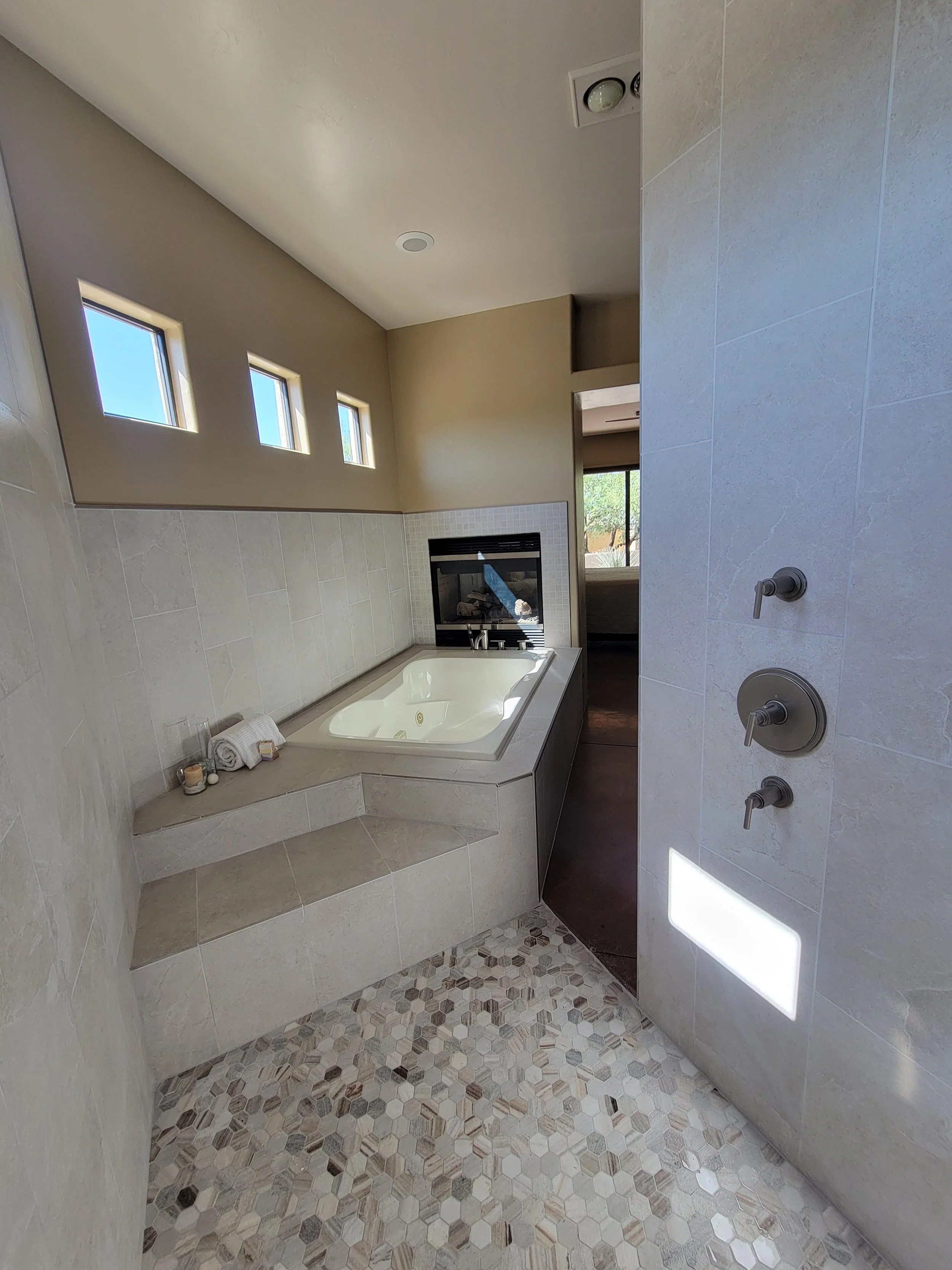 Bathroom with a bathtub, three small windows, a shower area with three controls, beige walls, tiled floors, and some rolled towels and candles near the bathtub.