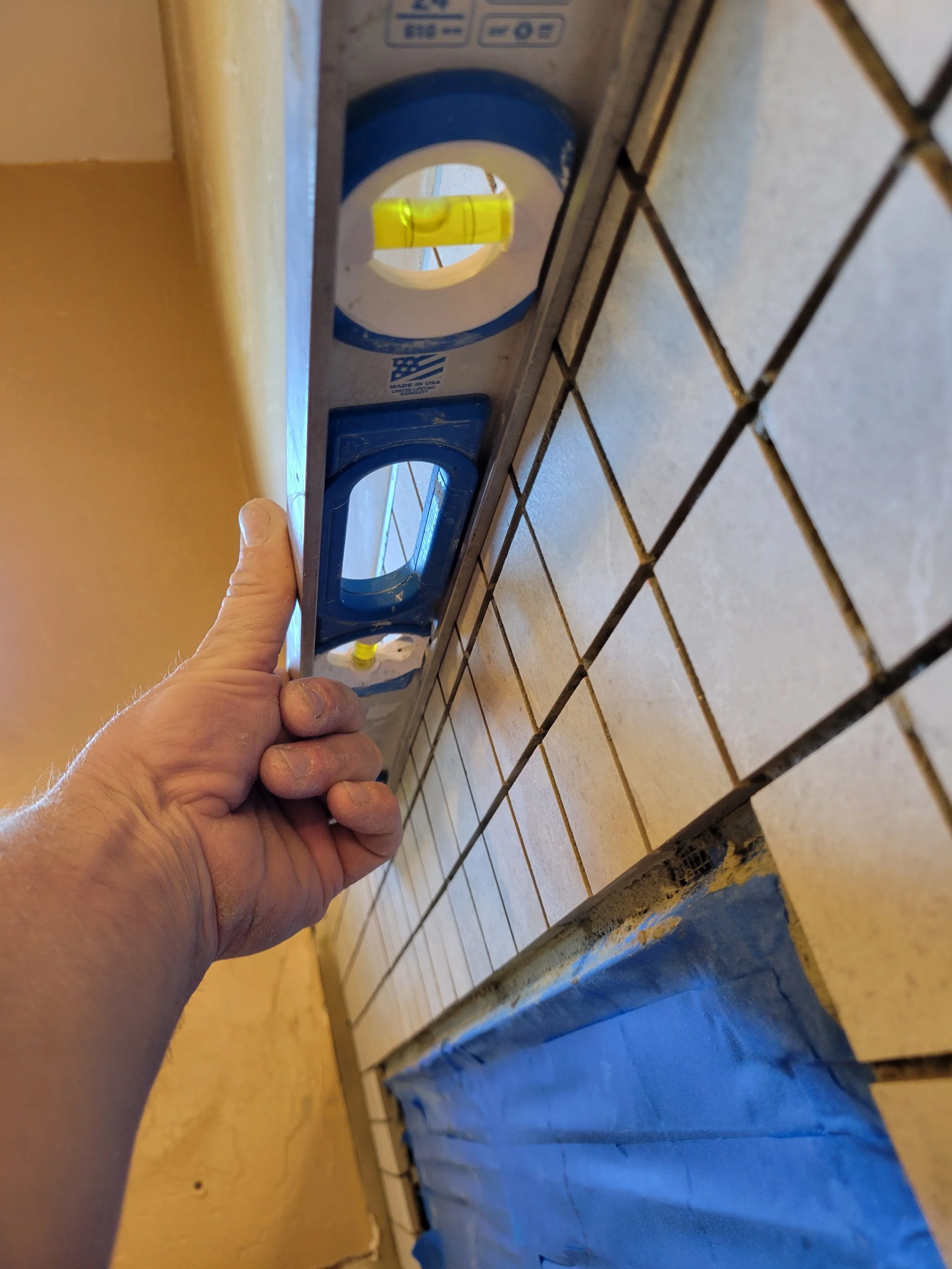 A person holds a blue level tool against a wall of tan-colored tiles to check their alignment. The tiles are small and square, and the level has a yellow bubble vial in the middle.