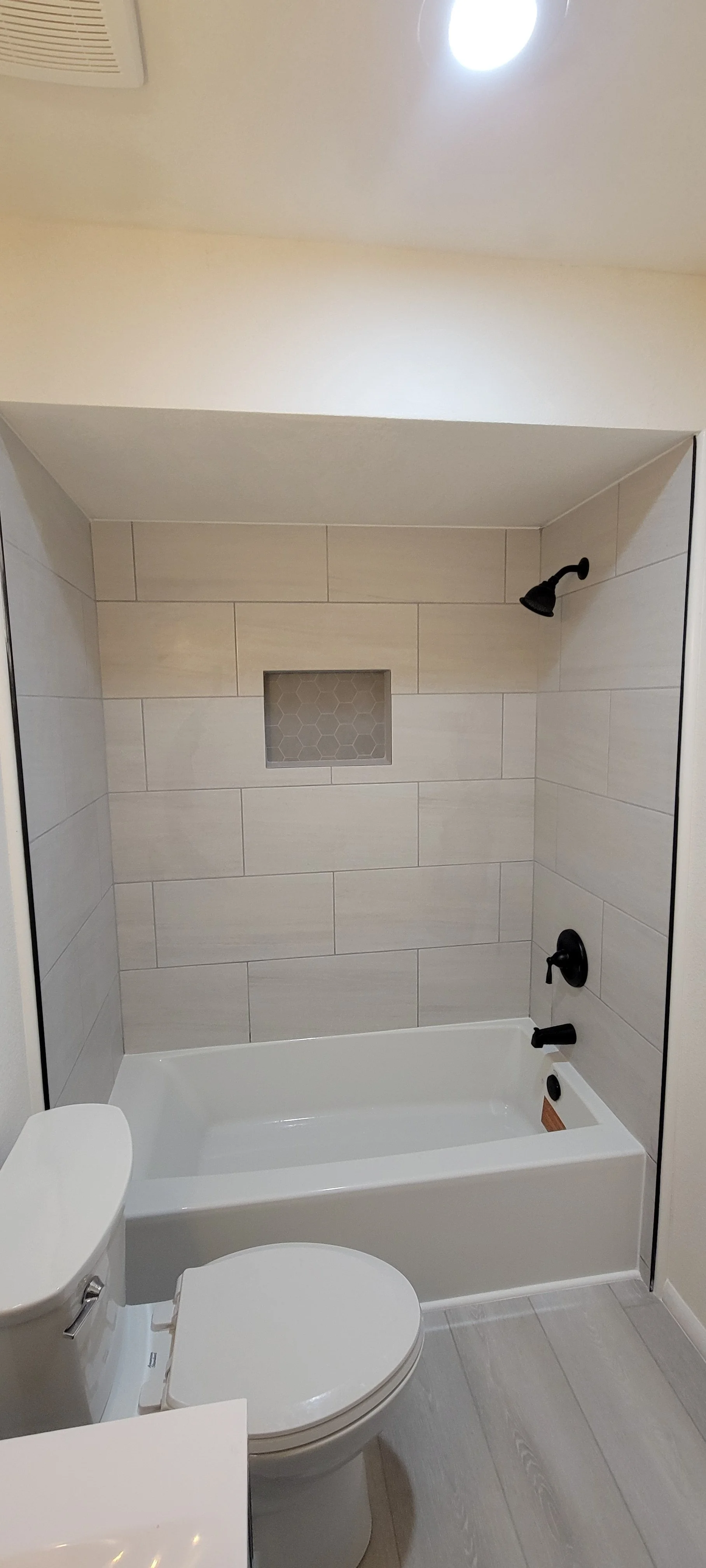 Bathroom with bathtub, toilet, beige tiled shower wall with built-in niche, black shower fixtures, and a ceiling light.