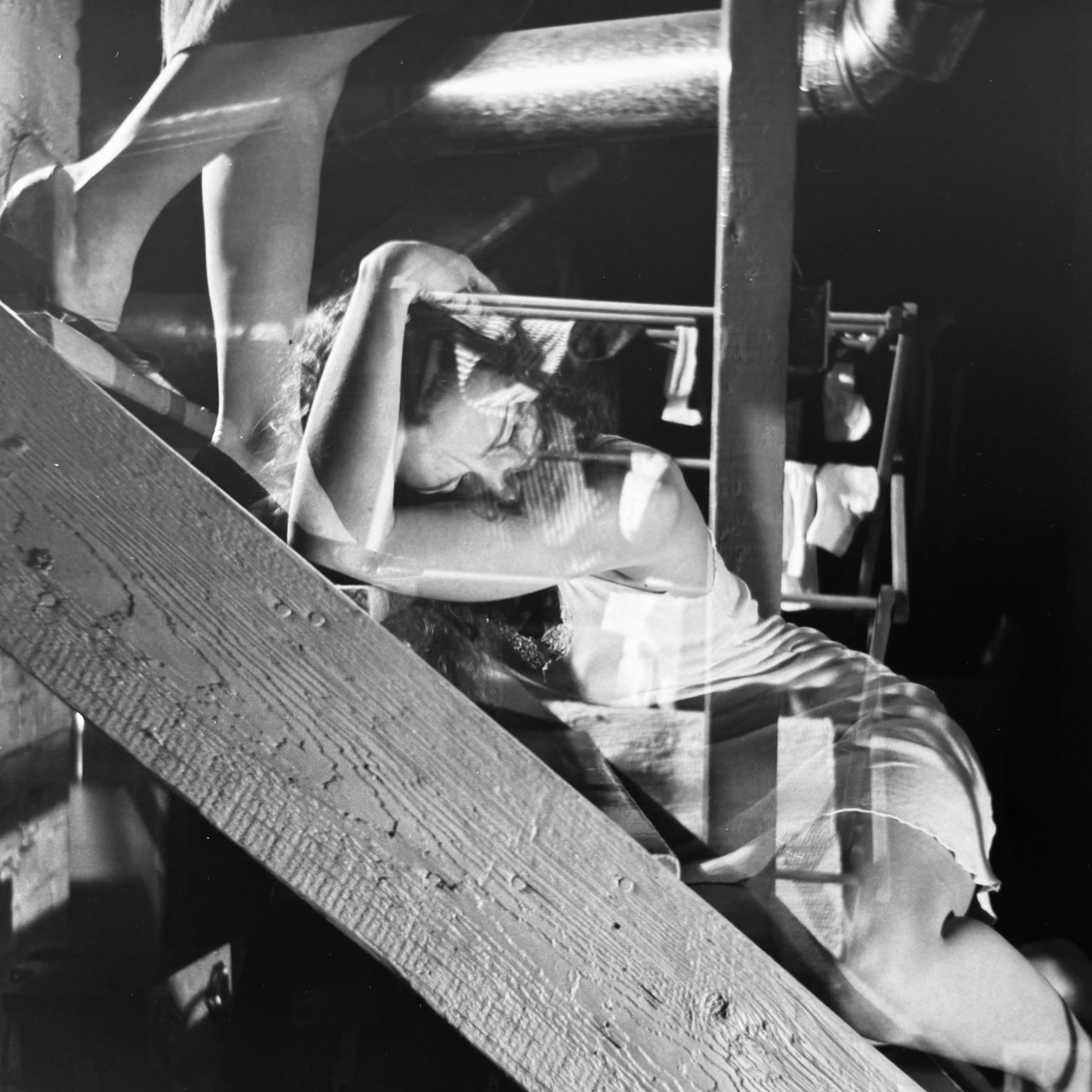 A black and white photograph of a person with long hair leaning on a wooden surface, underneath a structure with various objects, possibly in an outdoor setting.