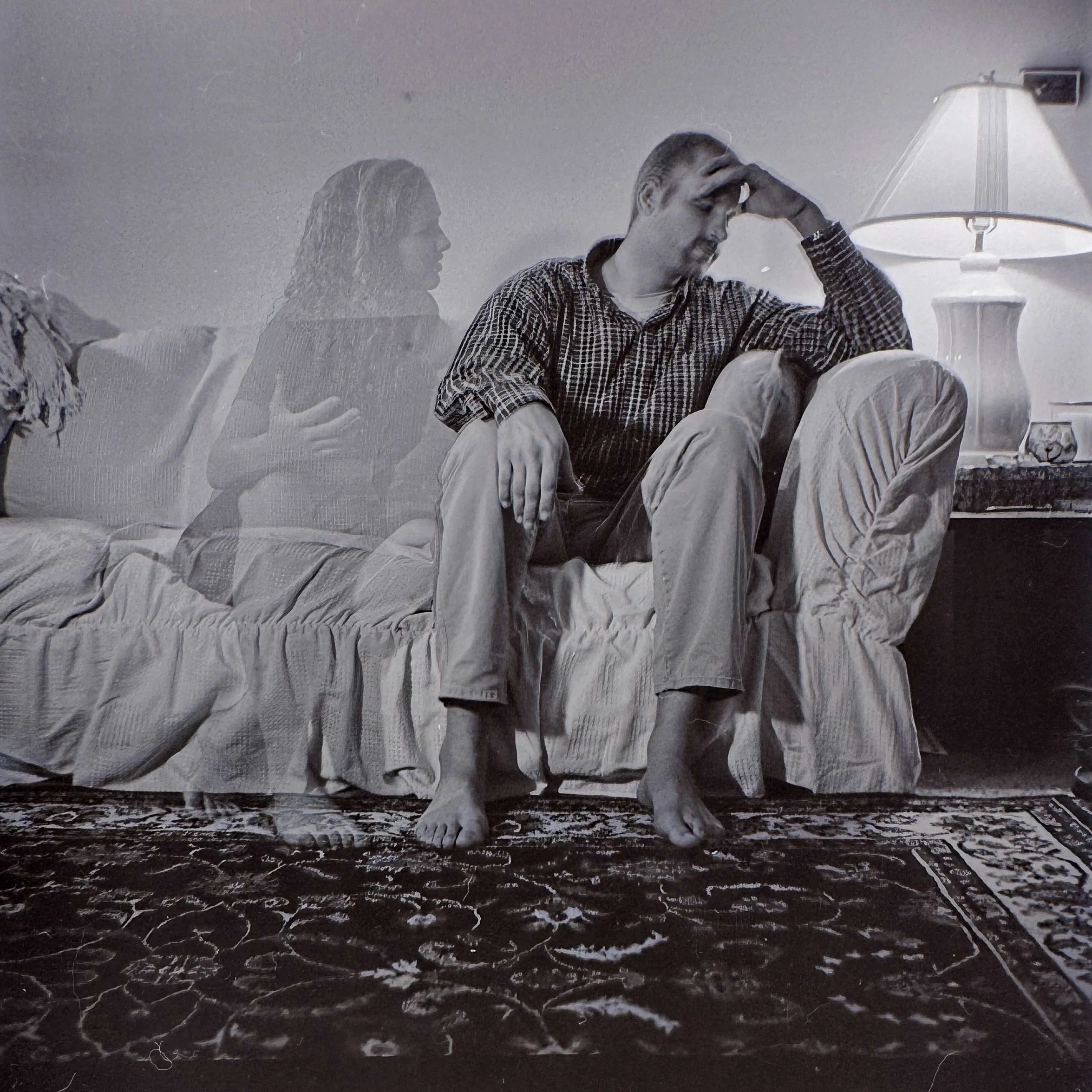 Black and white photo of two people sitting at a table, with the man on the right passing his hand over his forehead, and a woman on the left with long hair, in a room with a table lamp and some objects.