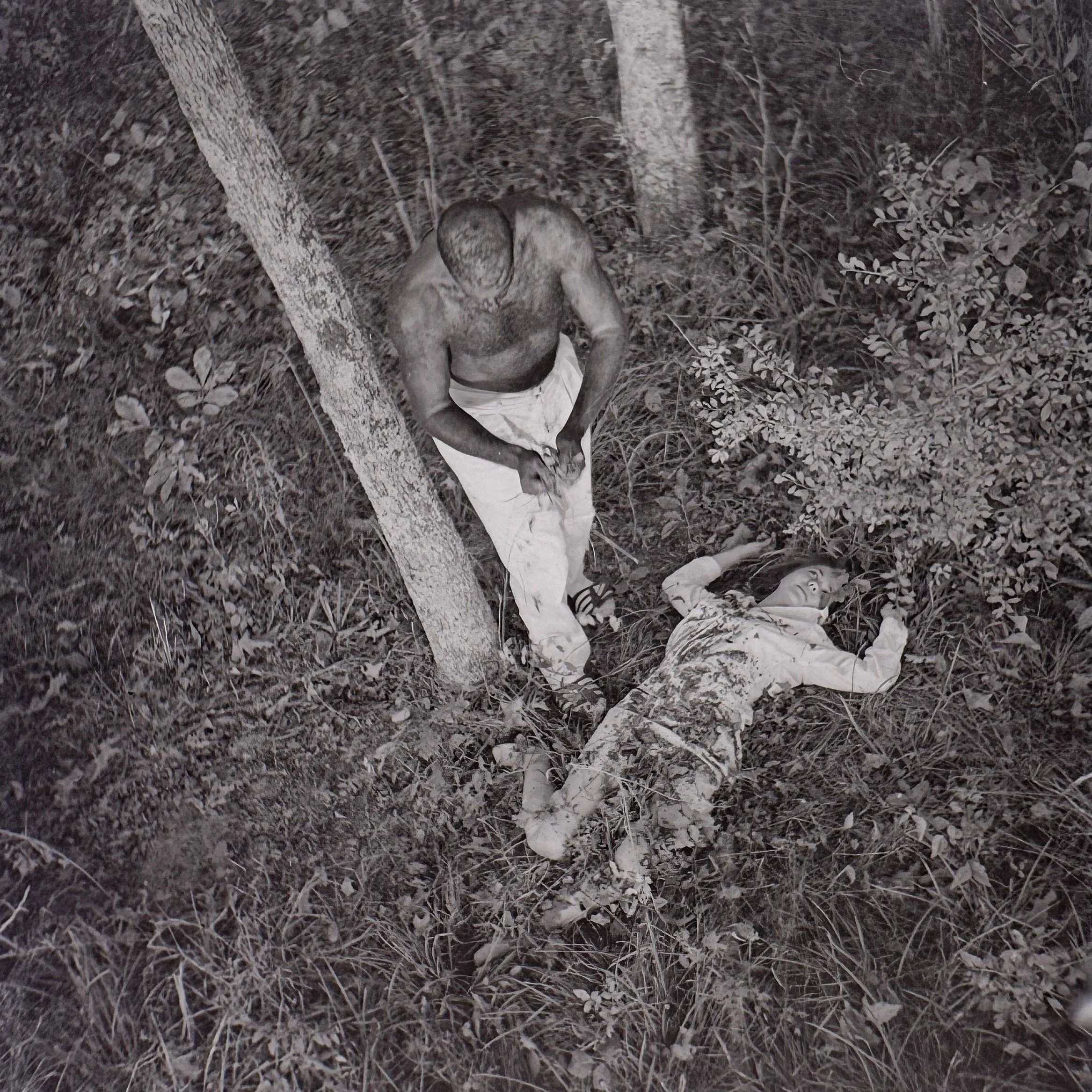 A person with dark hair, wearing a light-colored shirt and light pants, is sitting on the ground among grass and small plants, surrounded by trees, at night. The image appears to be in black and white.