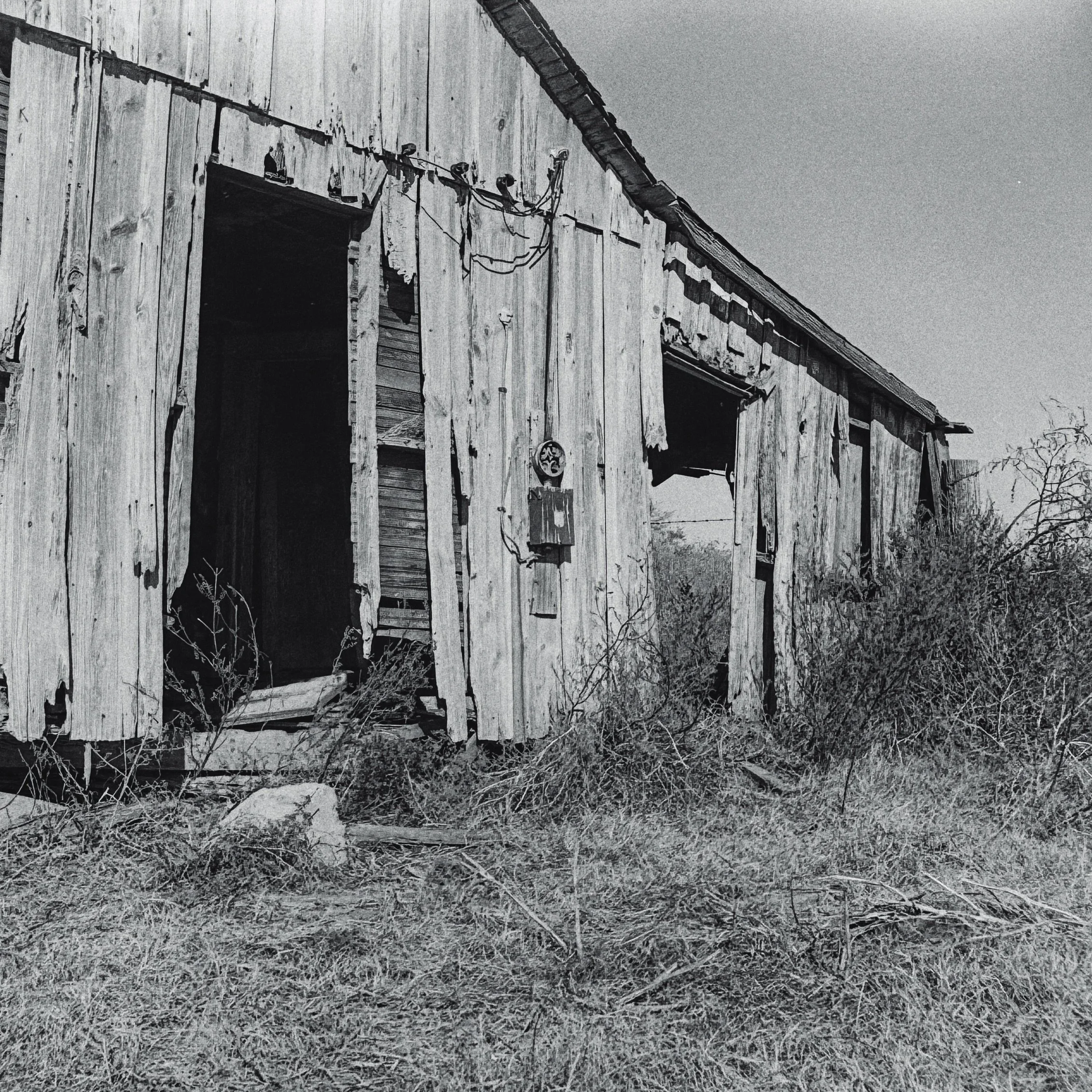 An old, weathered wooden structure with peeling paint and exposed boards, with an open doorway, wires hanging from the top, and sparse dry grass in the foreground. The sky above is cloudy.