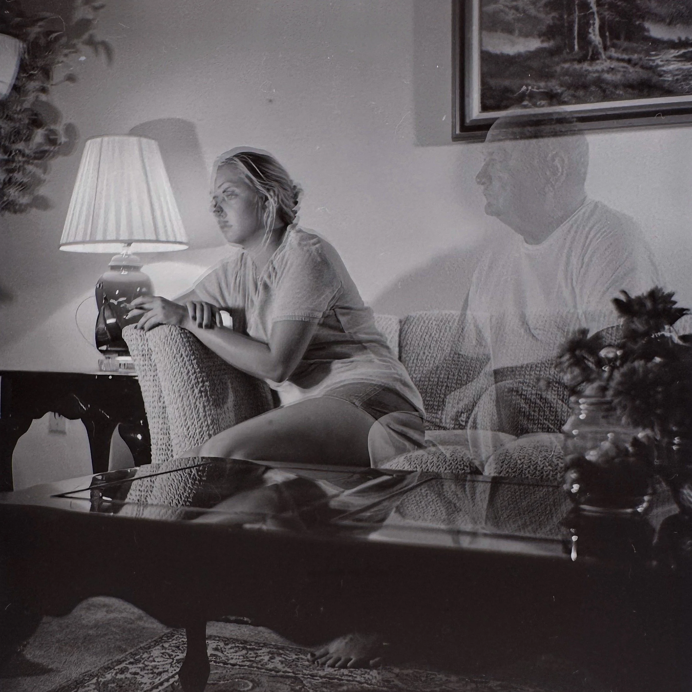 A grayscale photo of a woman and a man sitting on a couch near a wall with a framed picture and a lamp.