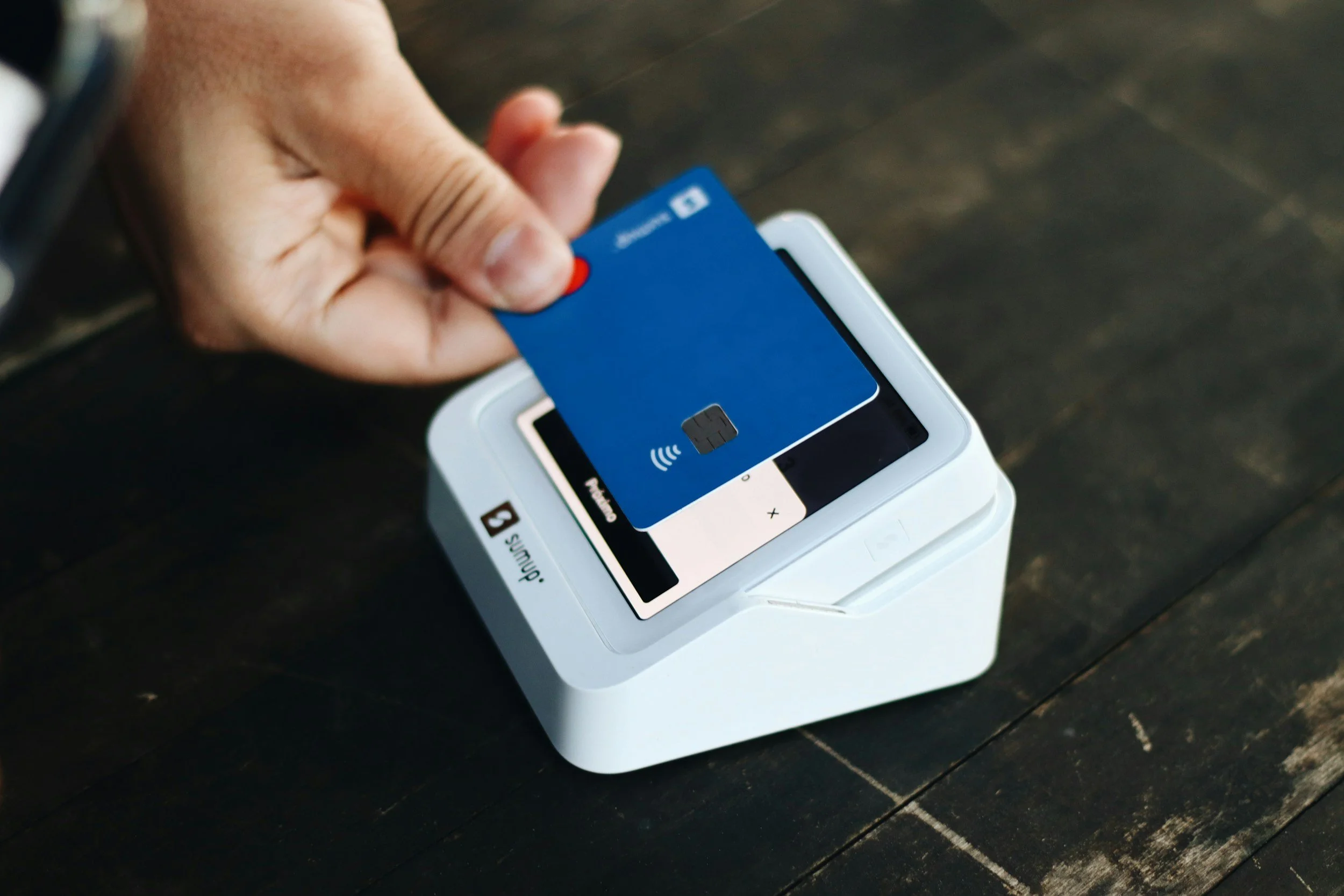 A person holding a blue credit card near a white contactless payment terminal on a dark wooden surface.