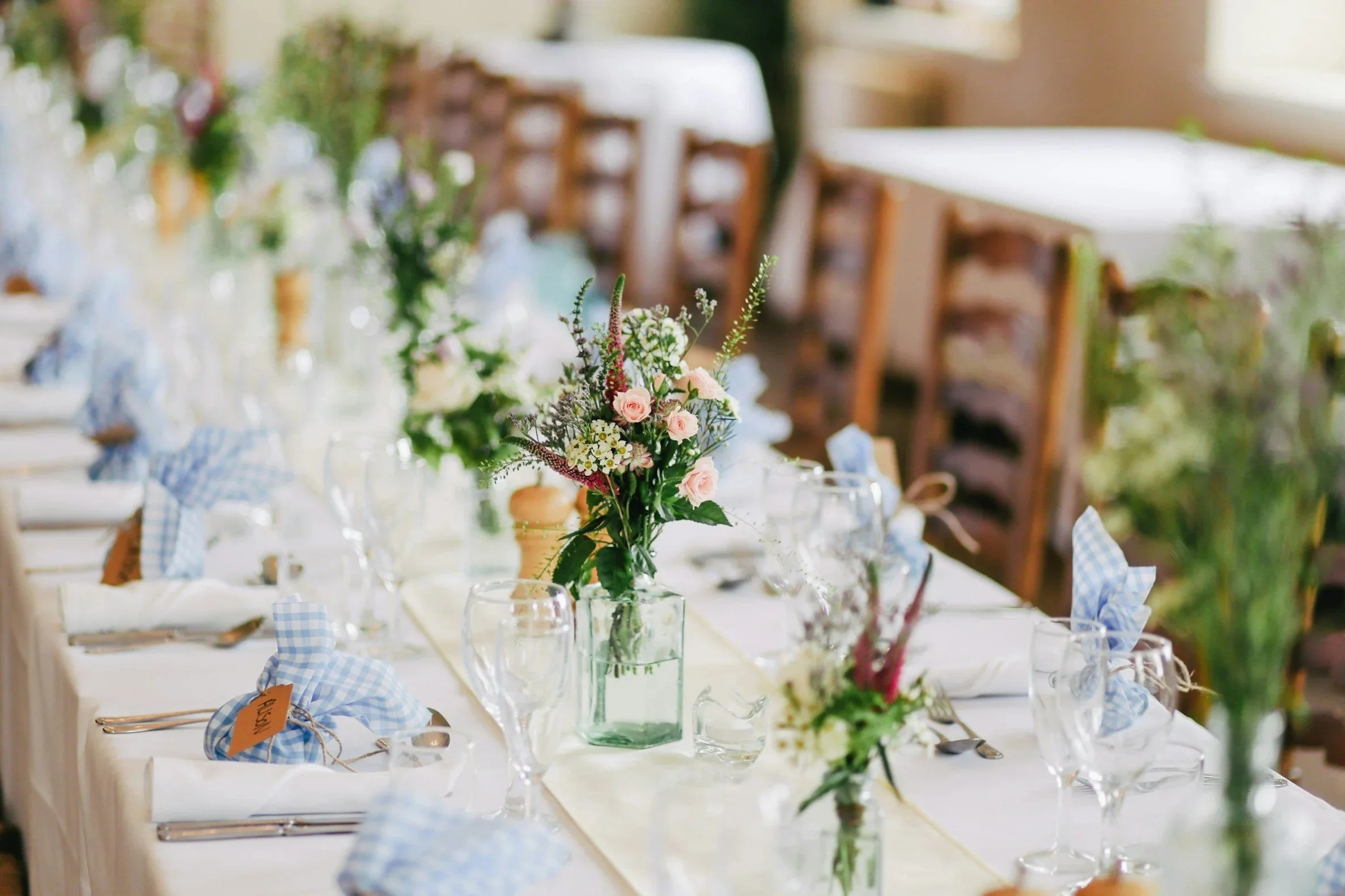 A banquet table decorated with a floral centerpiece featuring pink roses and white daisies, set with glassware, silverware, and blue checkered napkins in a bright, elegant setting.