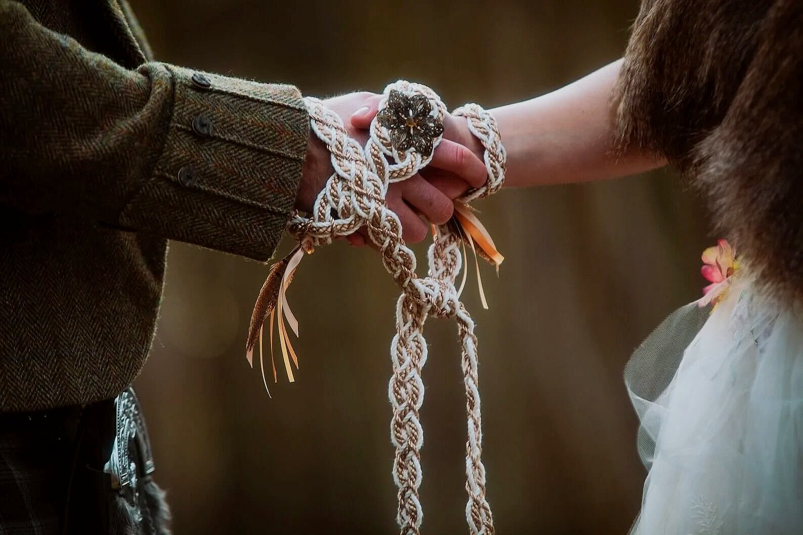 Two people shaking hands, one wearing a brown tweed jacket and the other a fluffy coat, with a decorative handmade rope bracelet on both their wrists.