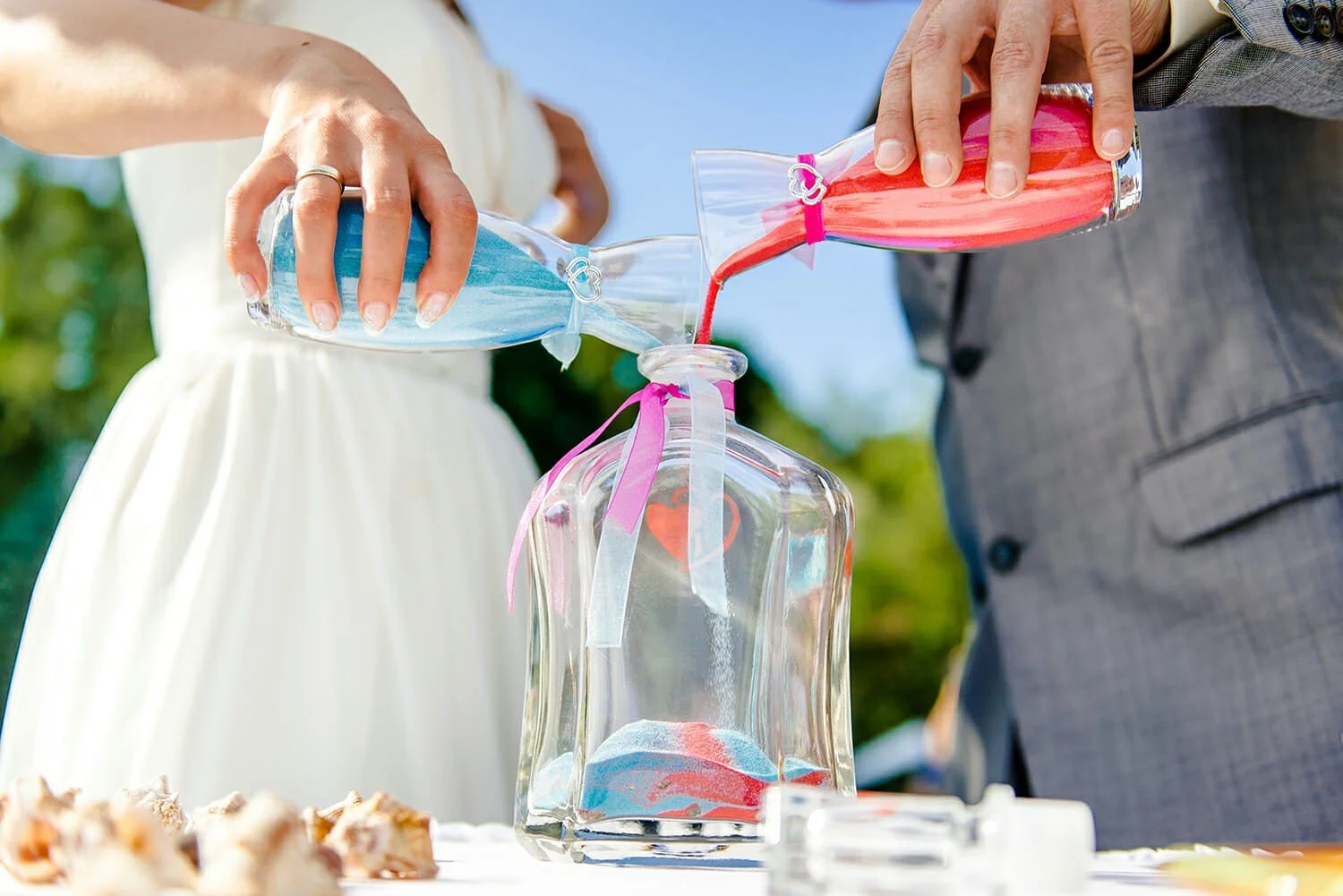 A couple is pouring pink and blue sand into a glass jar decorated with ribbons and a heart sticker during a wedding ceremony.