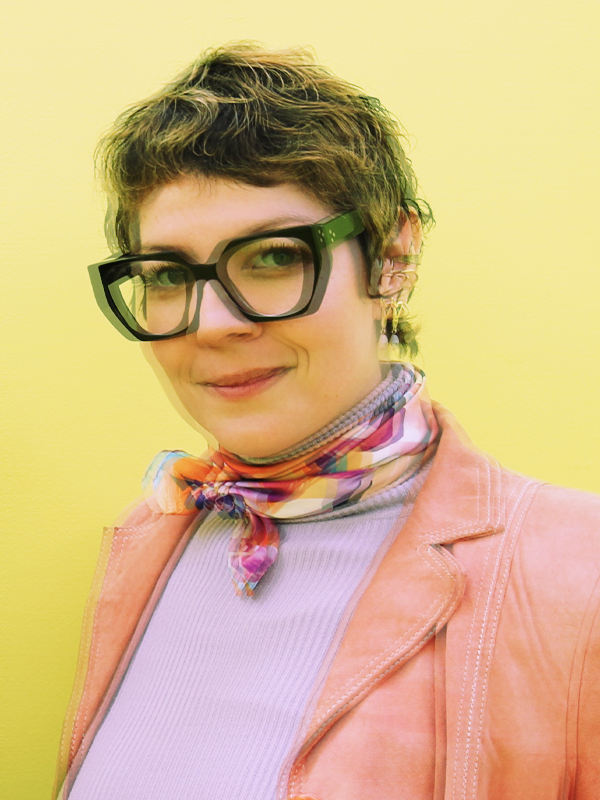 A woman with short brown hair wearing large black glasses, a pink jacket, a light purple top, and a multicolored scarf, smiling against a yellow background.