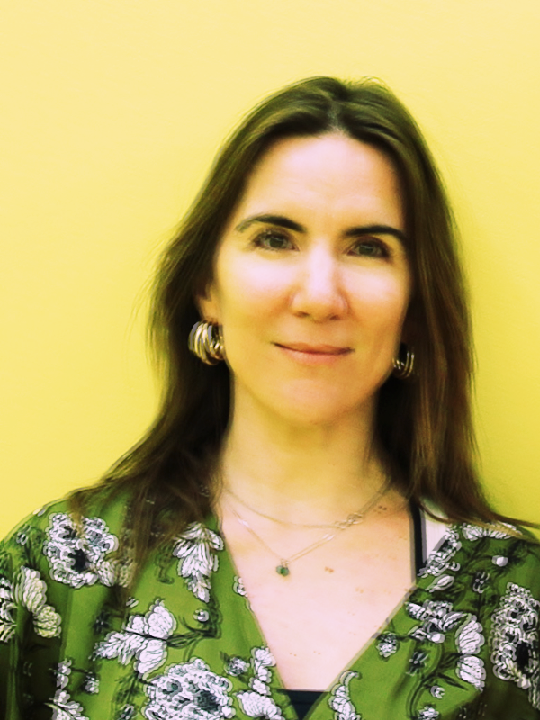 A woman with shoulder-length brown hair, wearing gold hoop earrings, a layered necklace, and a green floral blouse, standing against a yellow background.