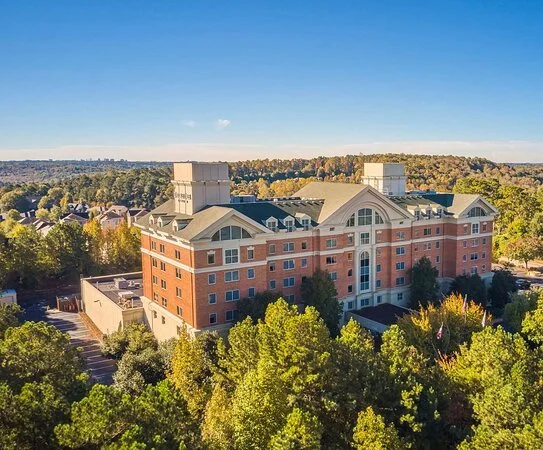 A large multi-story brick building surrounded by green trees under a clear blue sky.