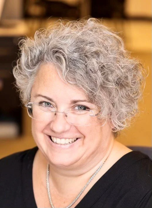 A smiling woman with gray, curly hair wearing glasses and a black top, sitting indoors.