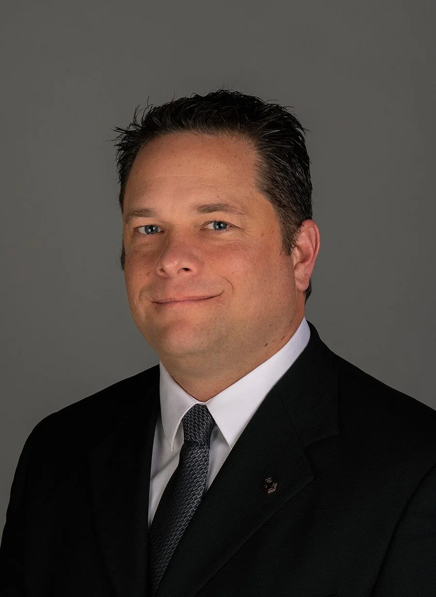 Portrait of a man with dark hair wearing a black suit, white shirt, and dark tie, against a gray background.