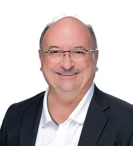 Portrait of a middle-aged man with glasses, smiling, wearing a dark blazer and white shirt, against a white background.